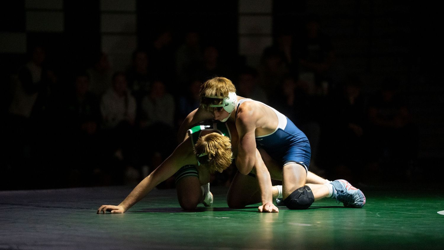 Blue singlet wrestler controls green singlet opponent in a takedown during a wrestling competition.