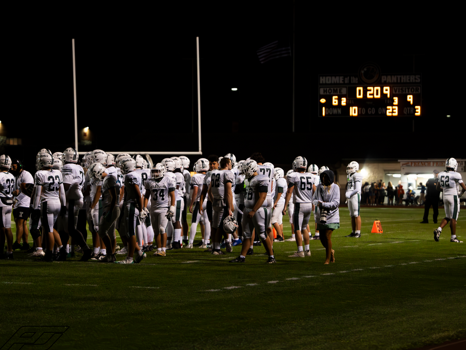 Football team stands on sideline with scoreboard showing 20-23 during night game.