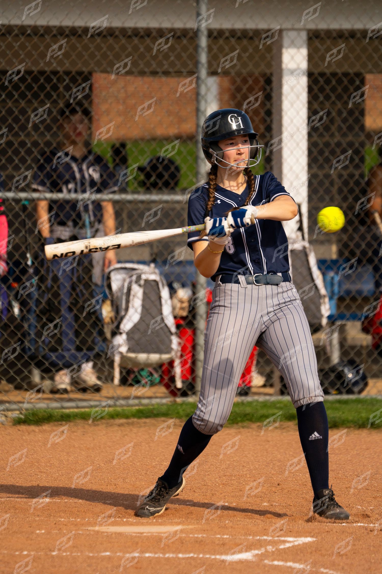 Female softball player in navy pinstripe uniform watches incoming yellow ball while preparing to swing bat.