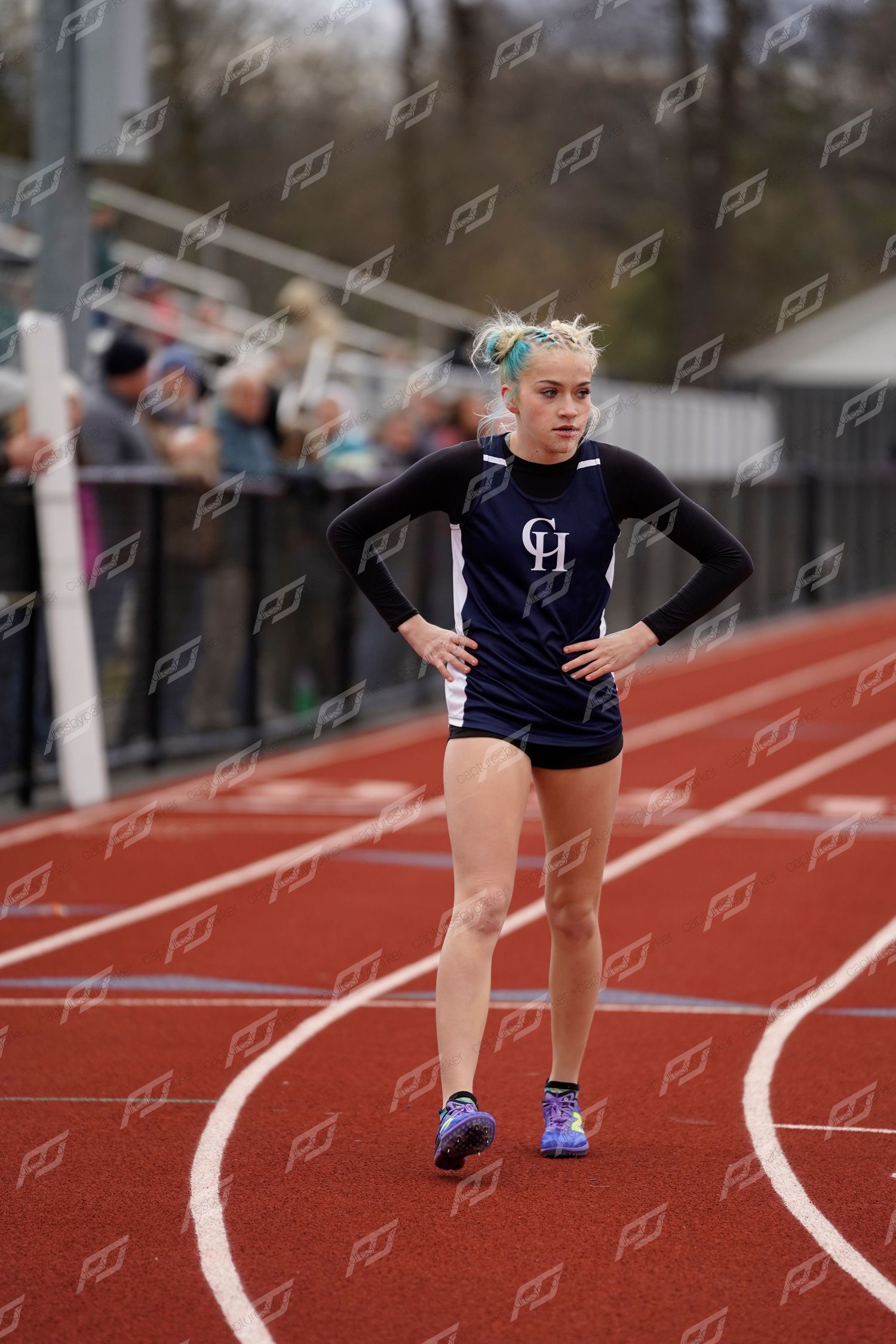 Female track athlete in navy CH uniform stands on red track with hands on hips between races.