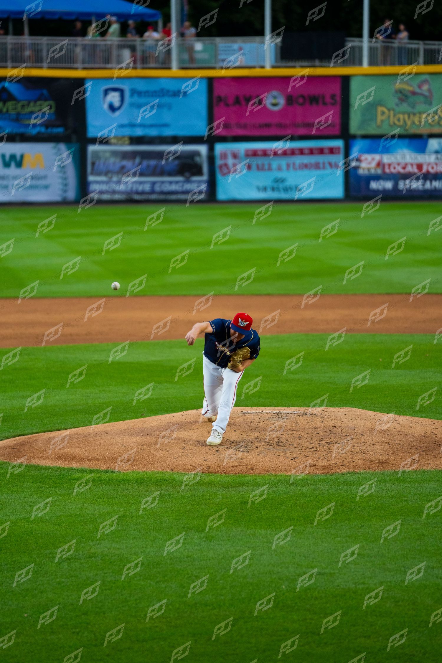 Minor league pitcher delivers pitch from mound in navy uniform during evening baseball game at stadium.