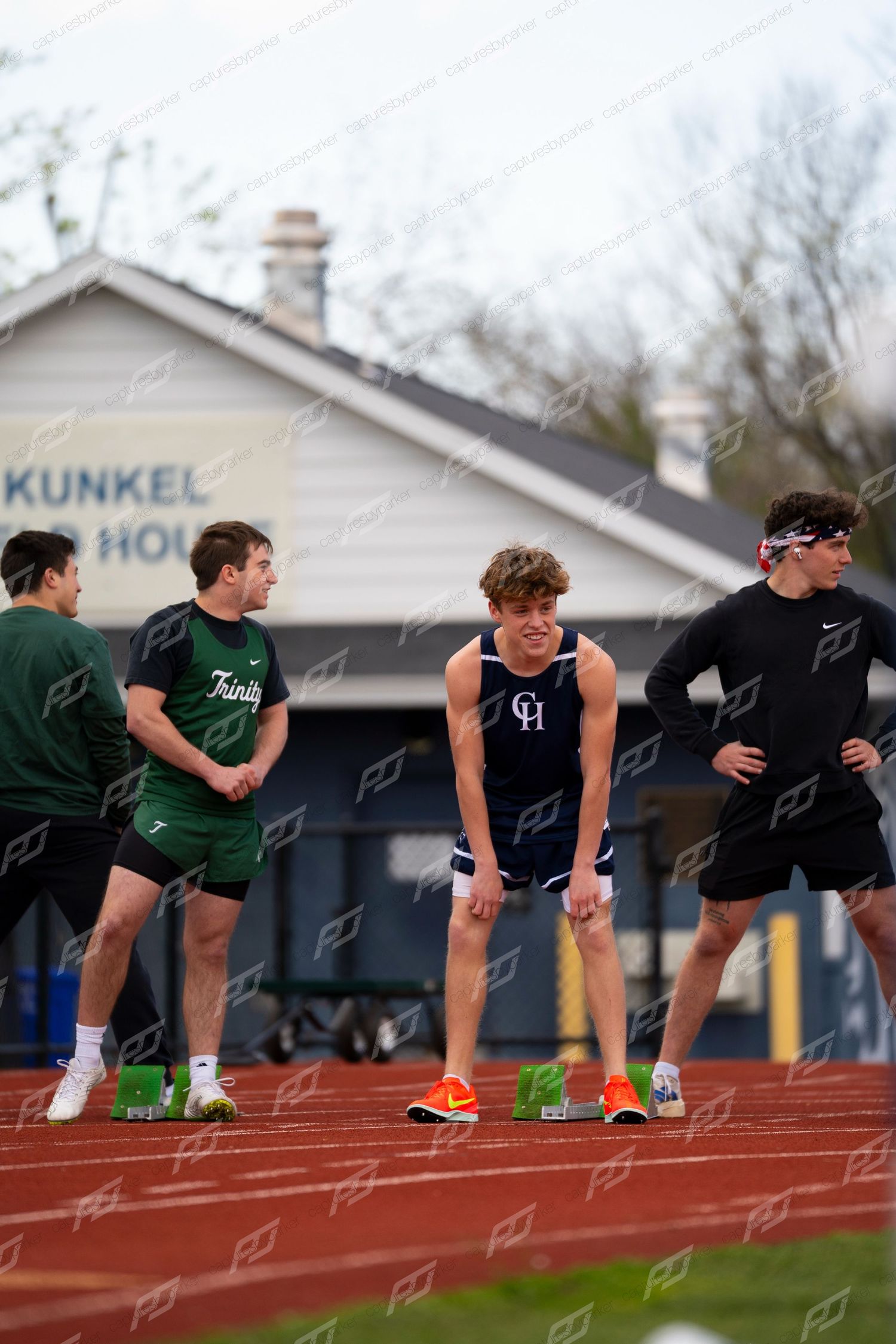 Male track athletes in blue and green uniforms prepare at starting blocks on red track.