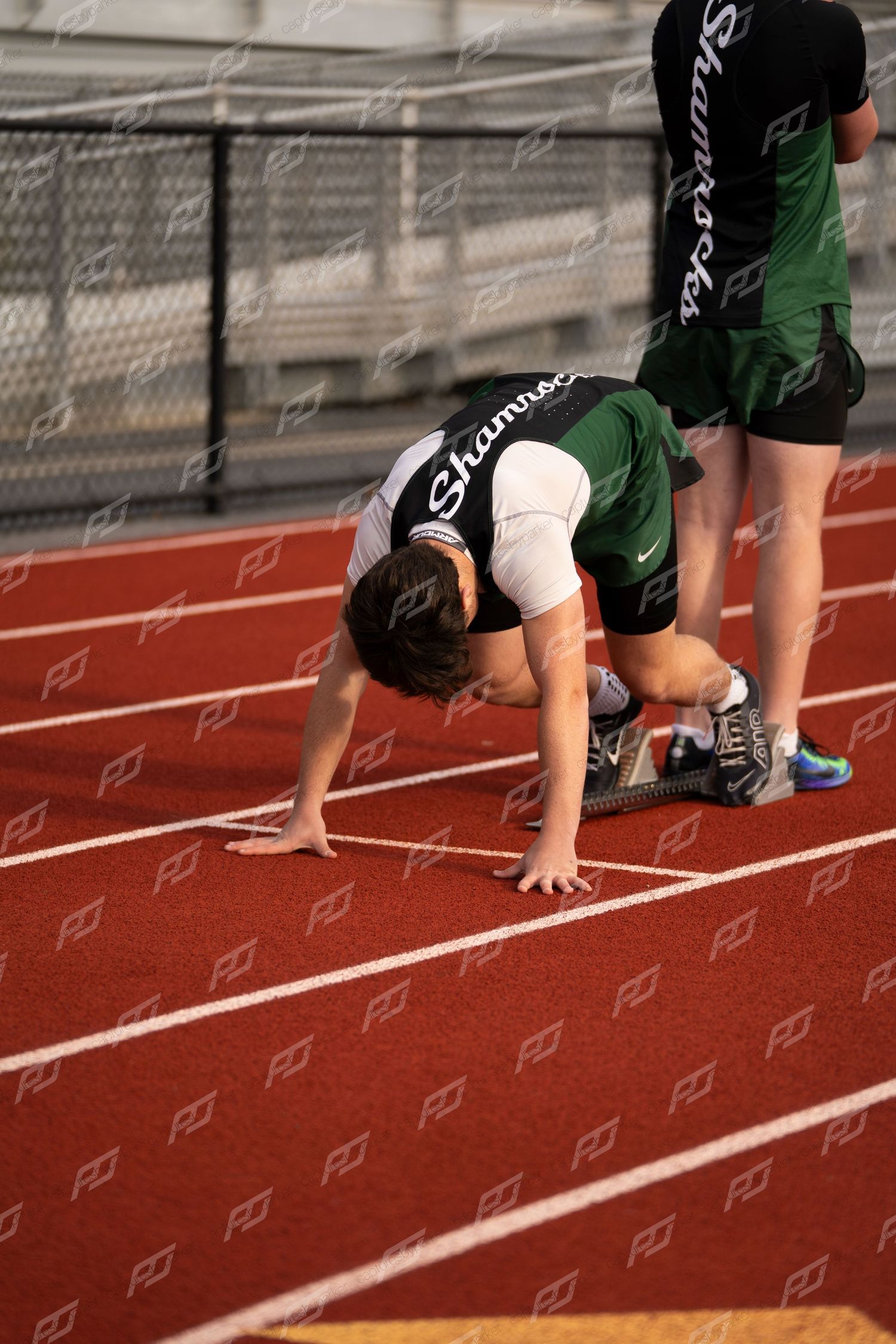 Young male runner in green Shamrocks jersey positioned in starting stance on red track.