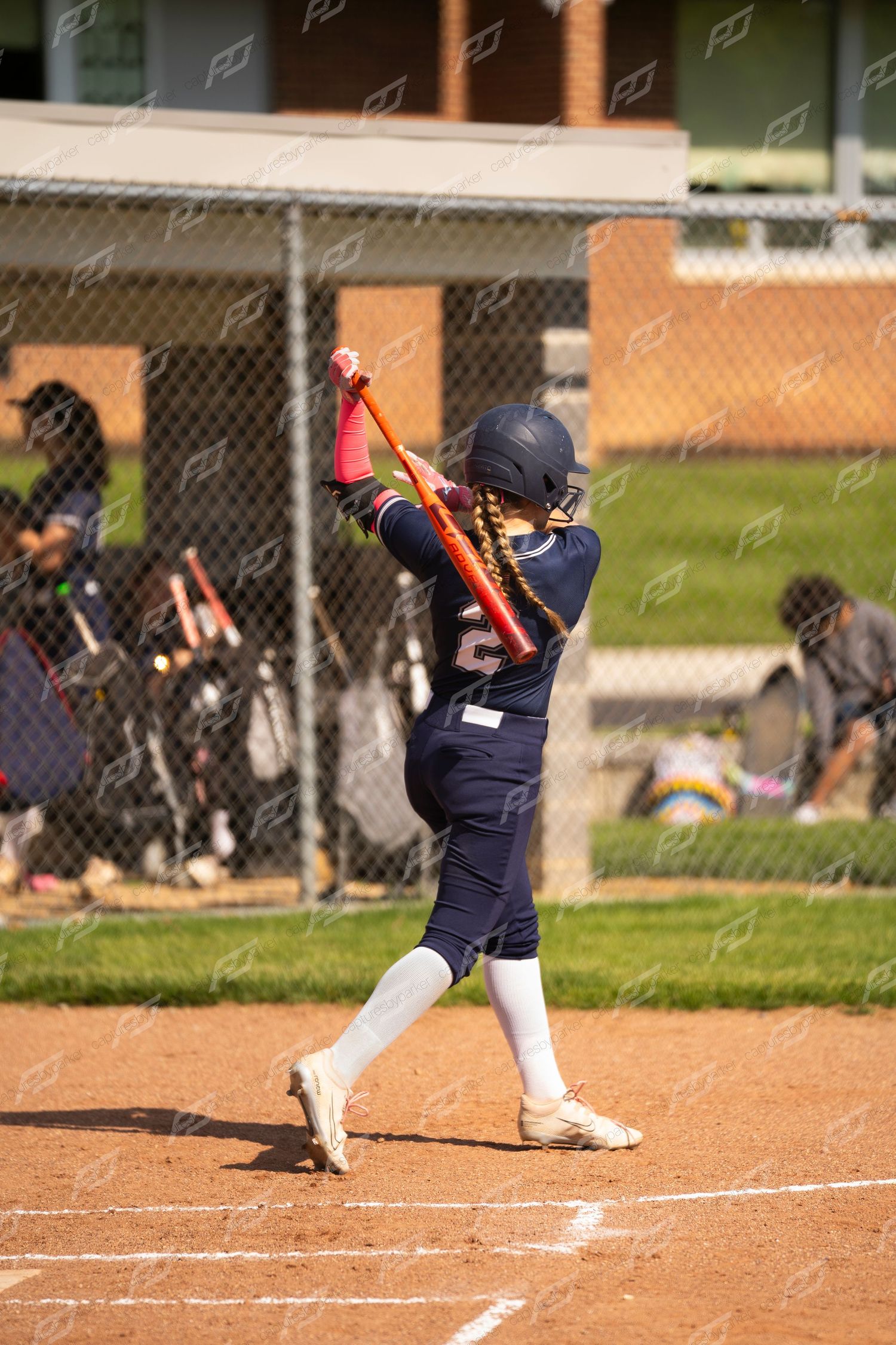 Young softball player in navy uniform follows through on swing with red bat at home plate.