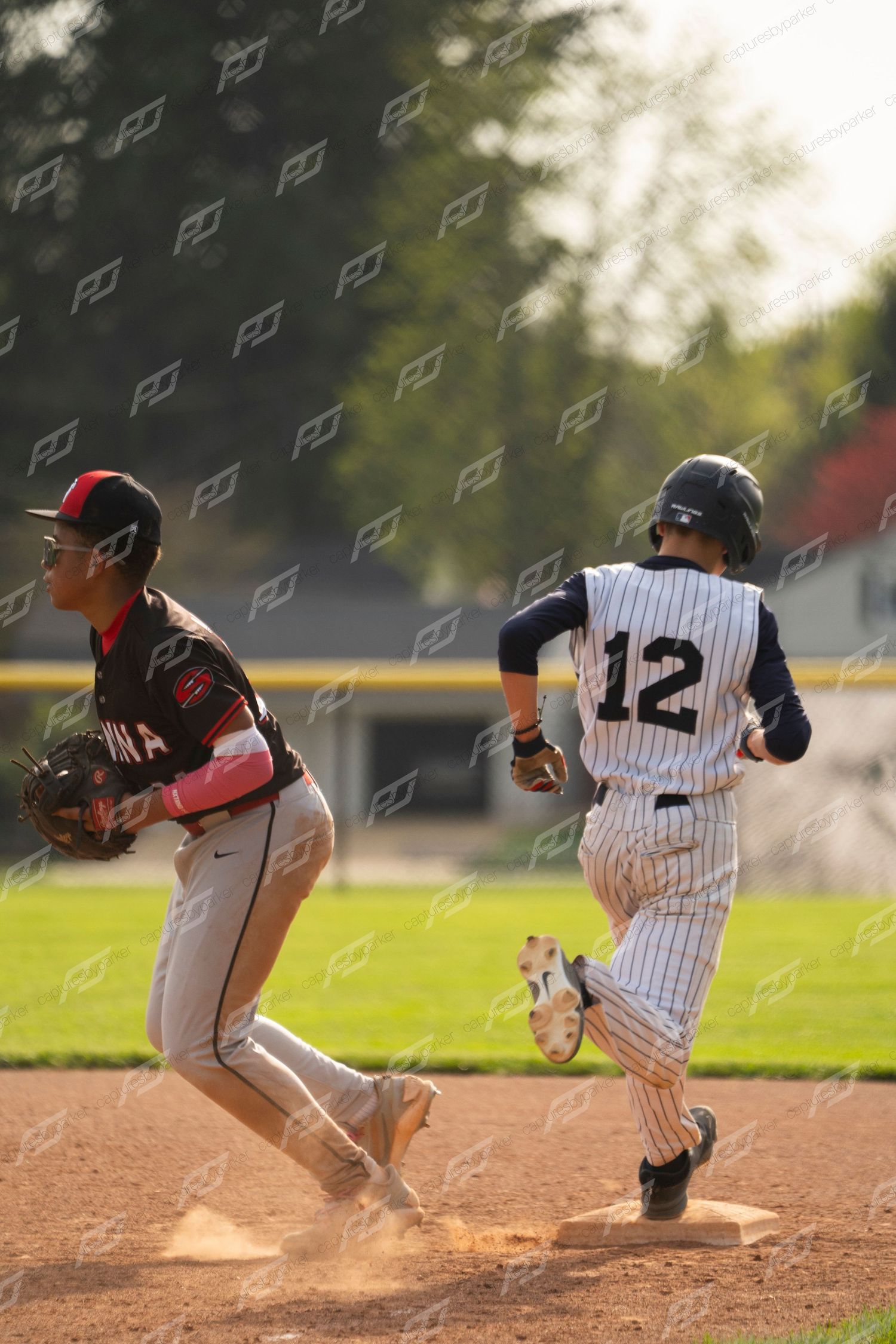 Baseball player number 12 in white pinstripes runs the bases as a fielder in black uniform moves nearby.