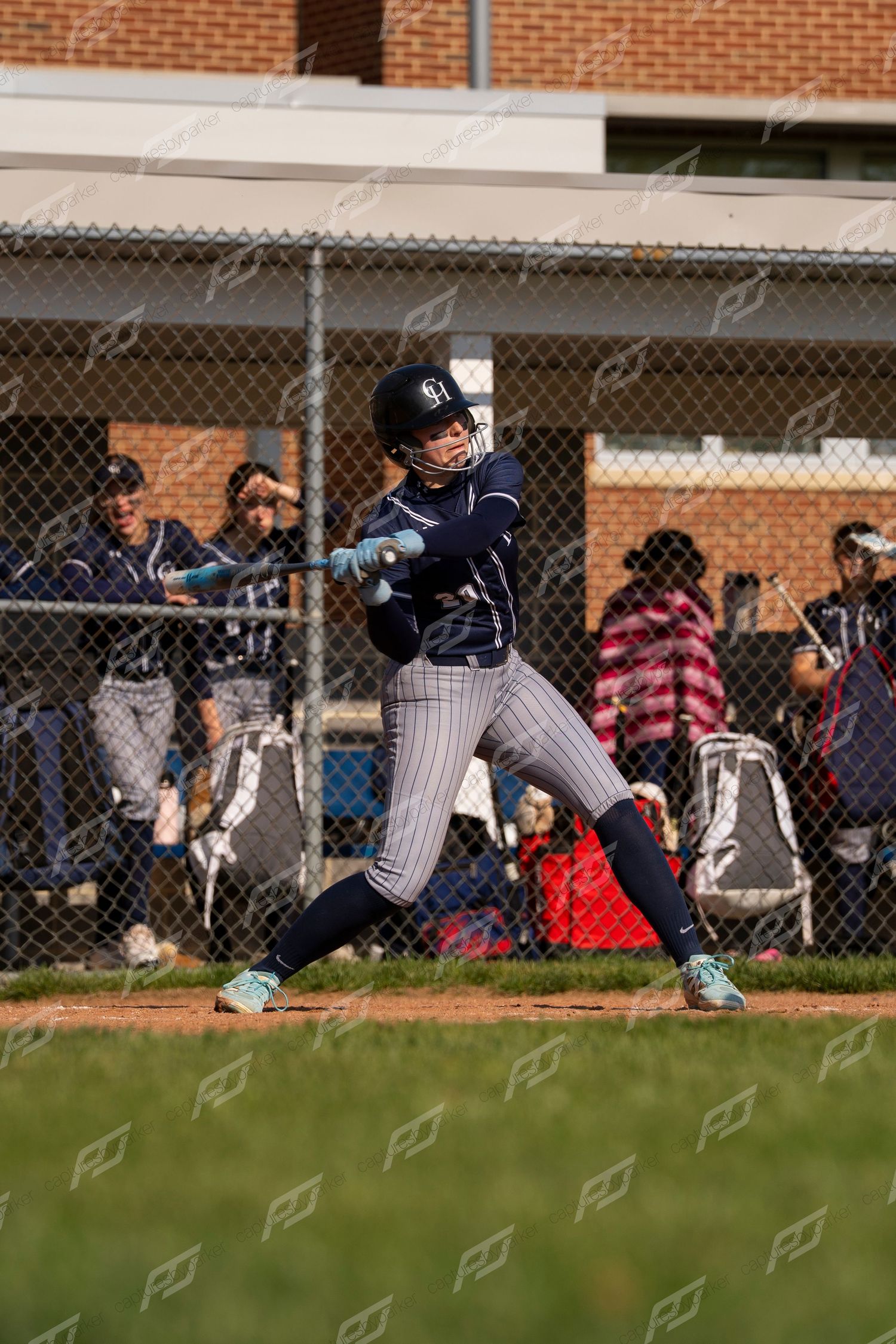 Youth softball player in navy pinstripe uniform swings bat at home plate with teammates watching from dugout behind fence.