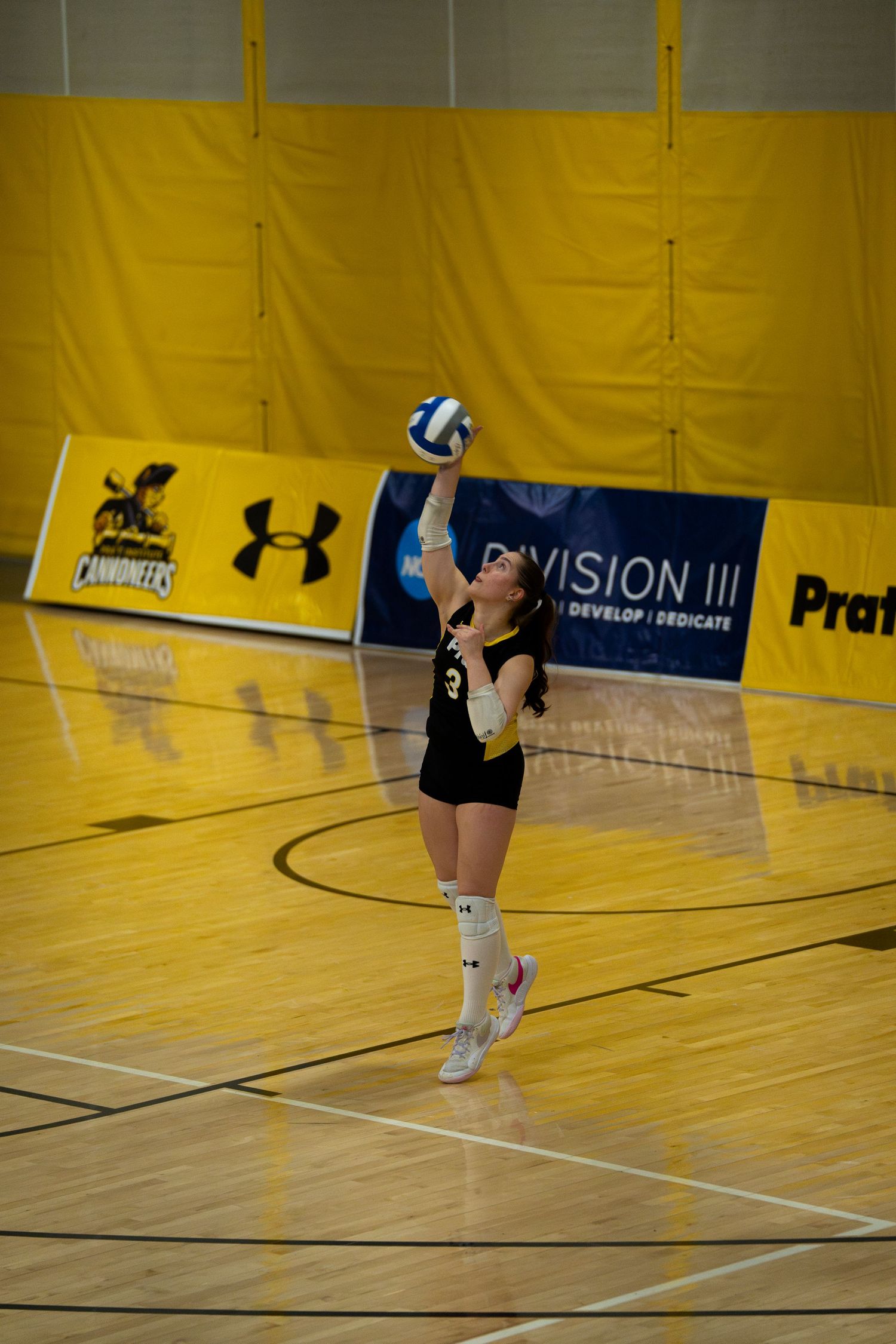 Female volleyball player in black uniform serving the ball during a Division III indoor match on a hardwood court.