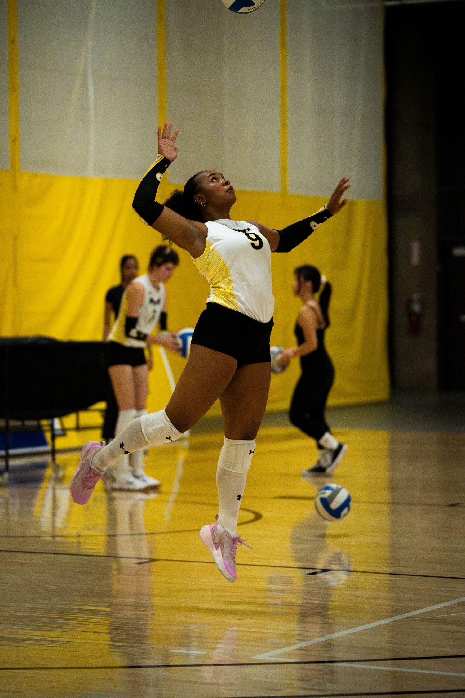 Female volleyball player in yellow and black uniform jumping to serve the ball during an indoor match.
