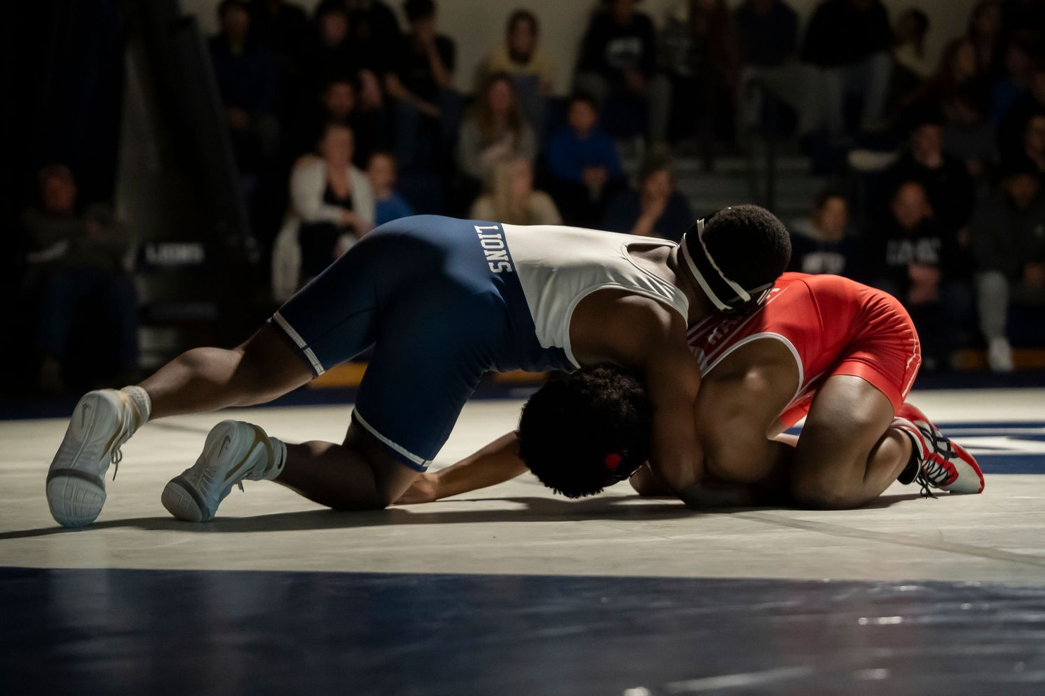 High school wrestlers grapple on a blue mat, one in navy blue singlet controlling opponent in red during match.