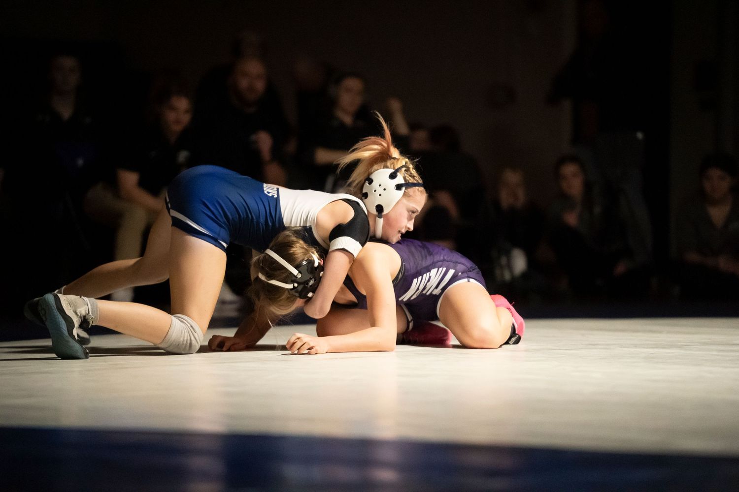 Female wrestler in blue singlet and headgear works to pin opponent on the wrestling mat.