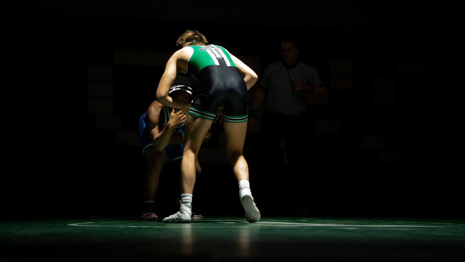 Two wrestlers compete under dramatic lighting, one in green singlet attempting a lift on opponent.
