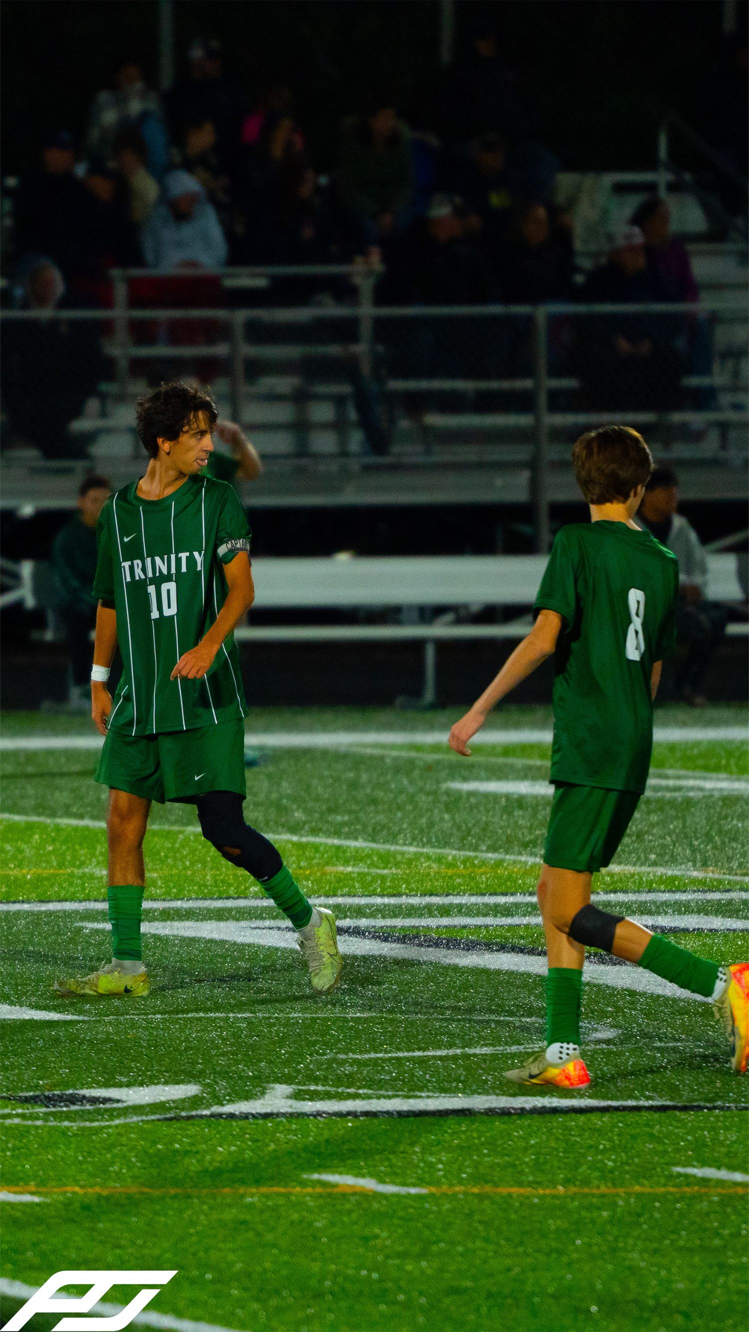 Trinity soccer players #10 and #8 in green uniforms on a wet turf field during a night game.