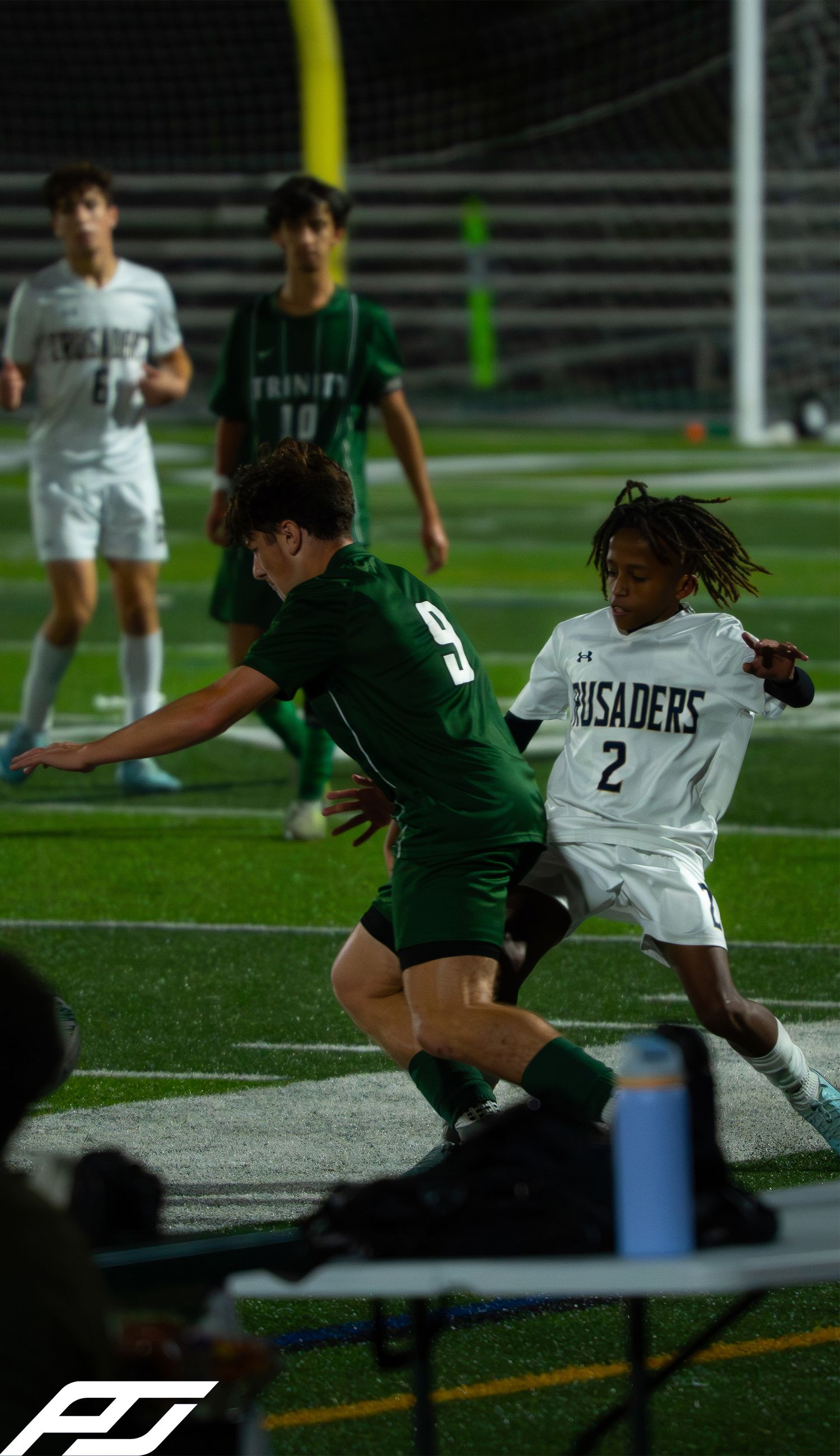 Soccer player in green Trinity jersey battles for the ball against Crusaders number 2 at night.