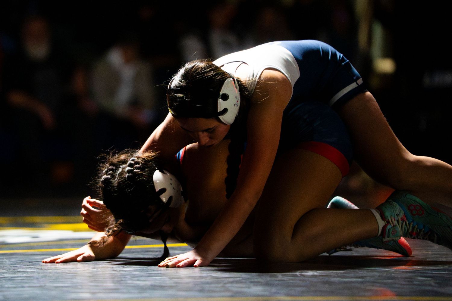 Two female wrestlers in singlets and headgear compete in a ground position during a wrestling match.