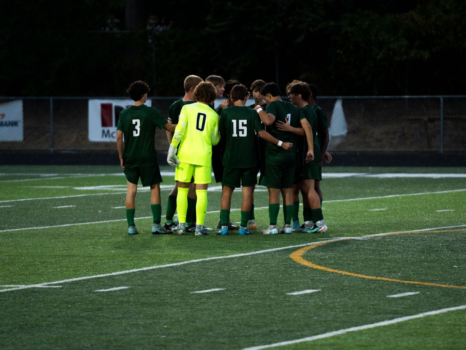Youth soccer team in green uniforms huddles together on artificial turf during a game.