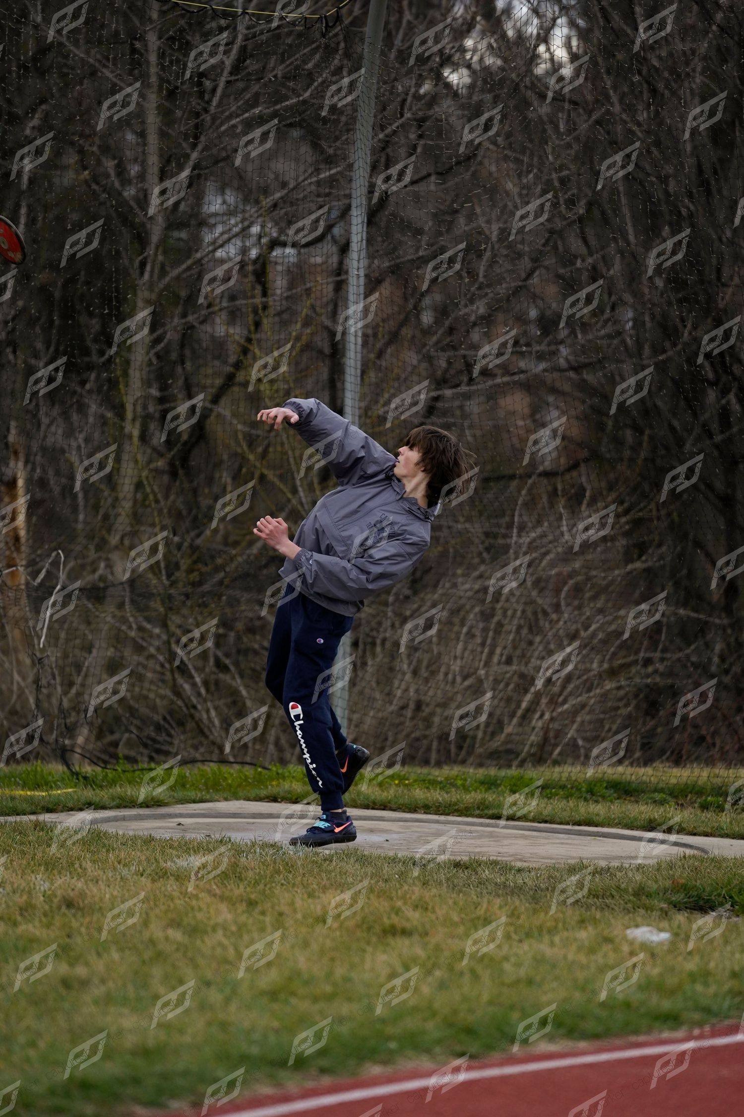 Athlete performs a discus throw on an outdoor track field surrounded by bare trees during a track and field meet.