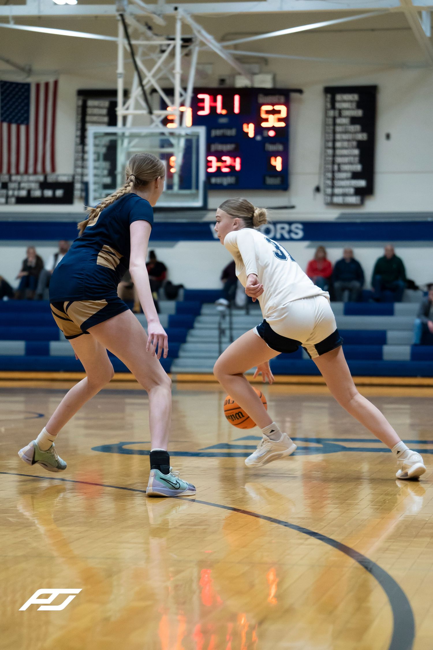 Female Lions basketball player dribbles against a defender with the scoreboard visible during a high school game.