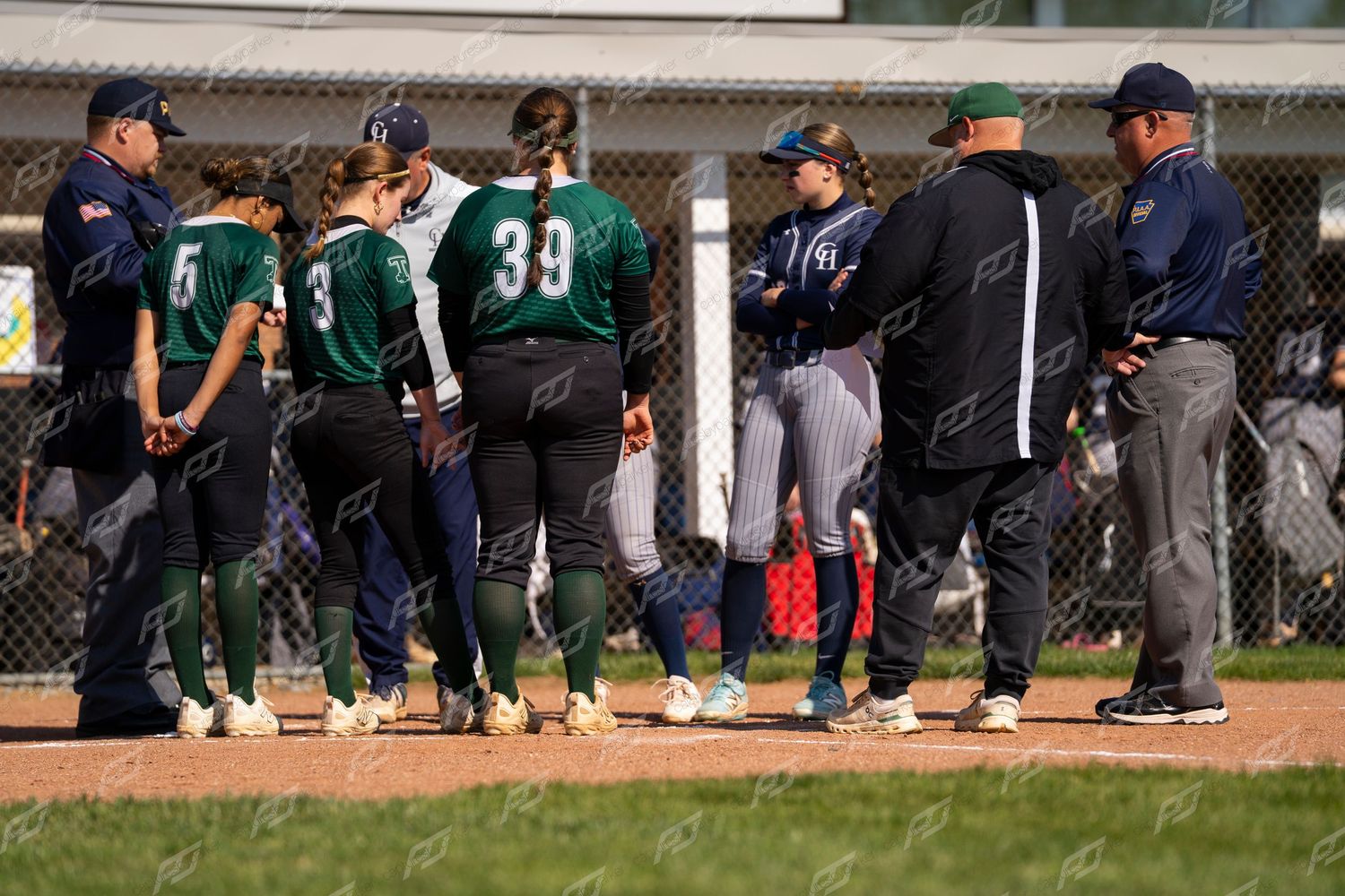 Softball players and coaches in navy and green uniforms gather on field during game timeout.