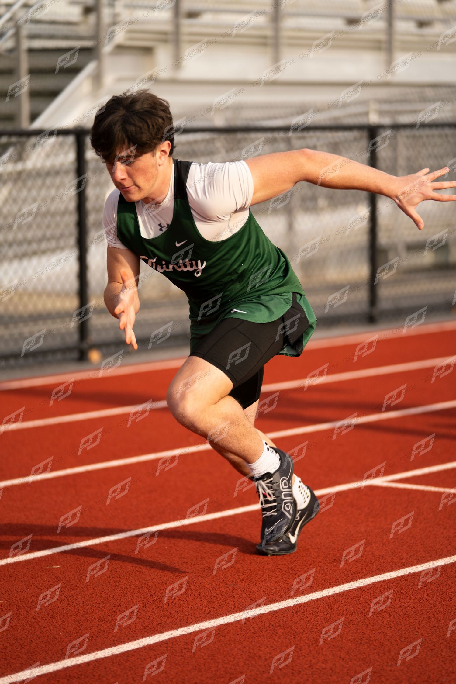 High school sprinter in green Trinity uniform drives powerfully out of starting blocks on track.