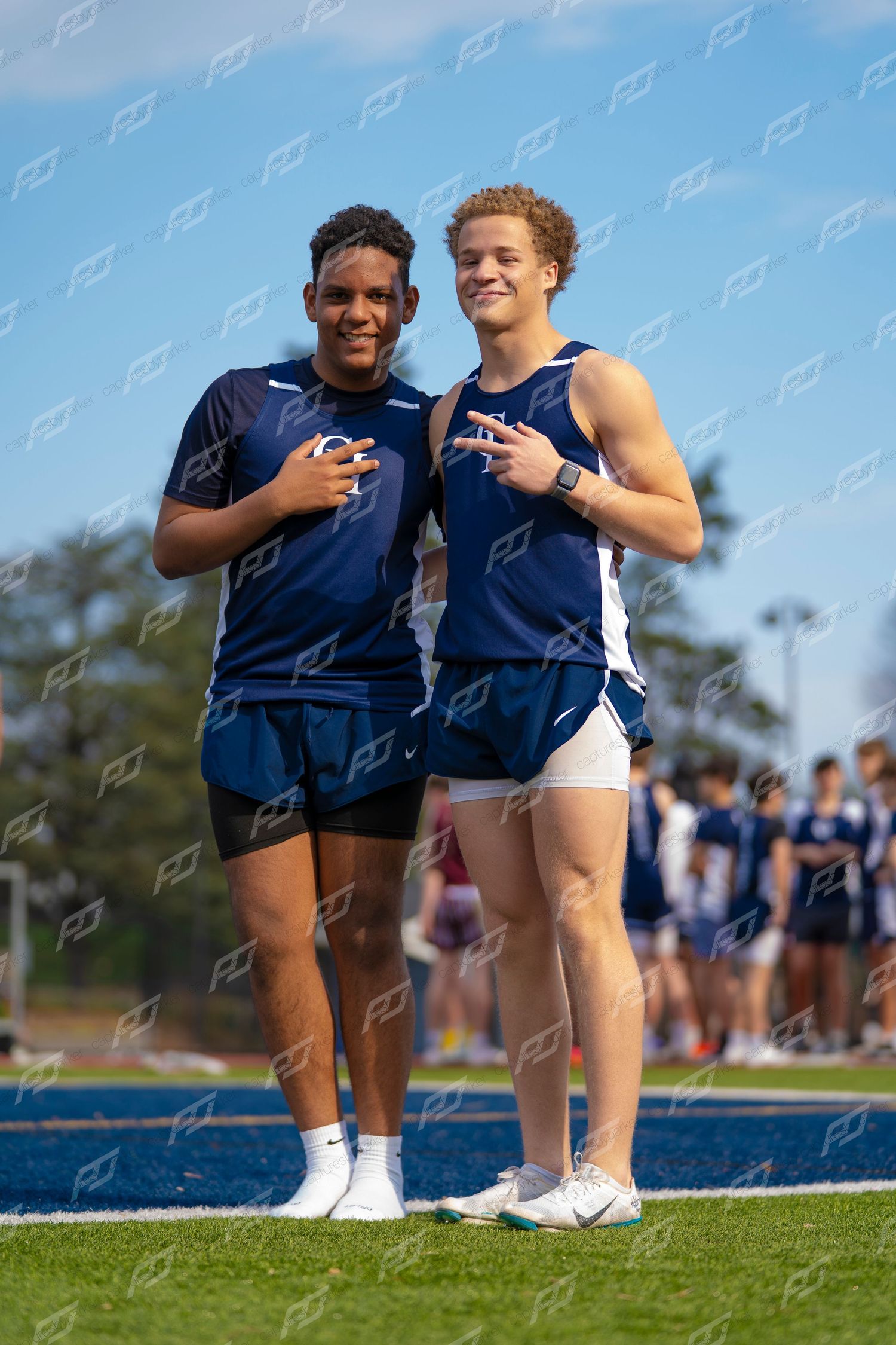 Two male track athletes in navy uniforms posing together on blue track field.