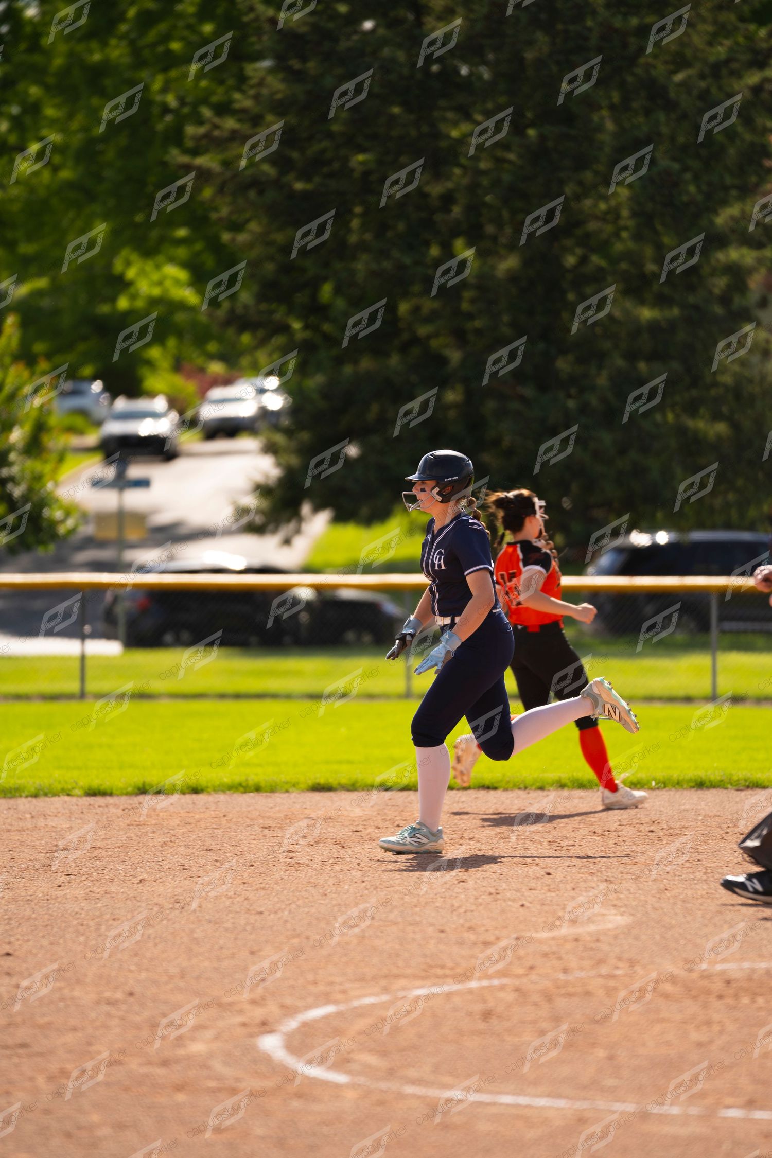 Softball player in navy uniform rounds a base ahead of an opponent in red during a game.
