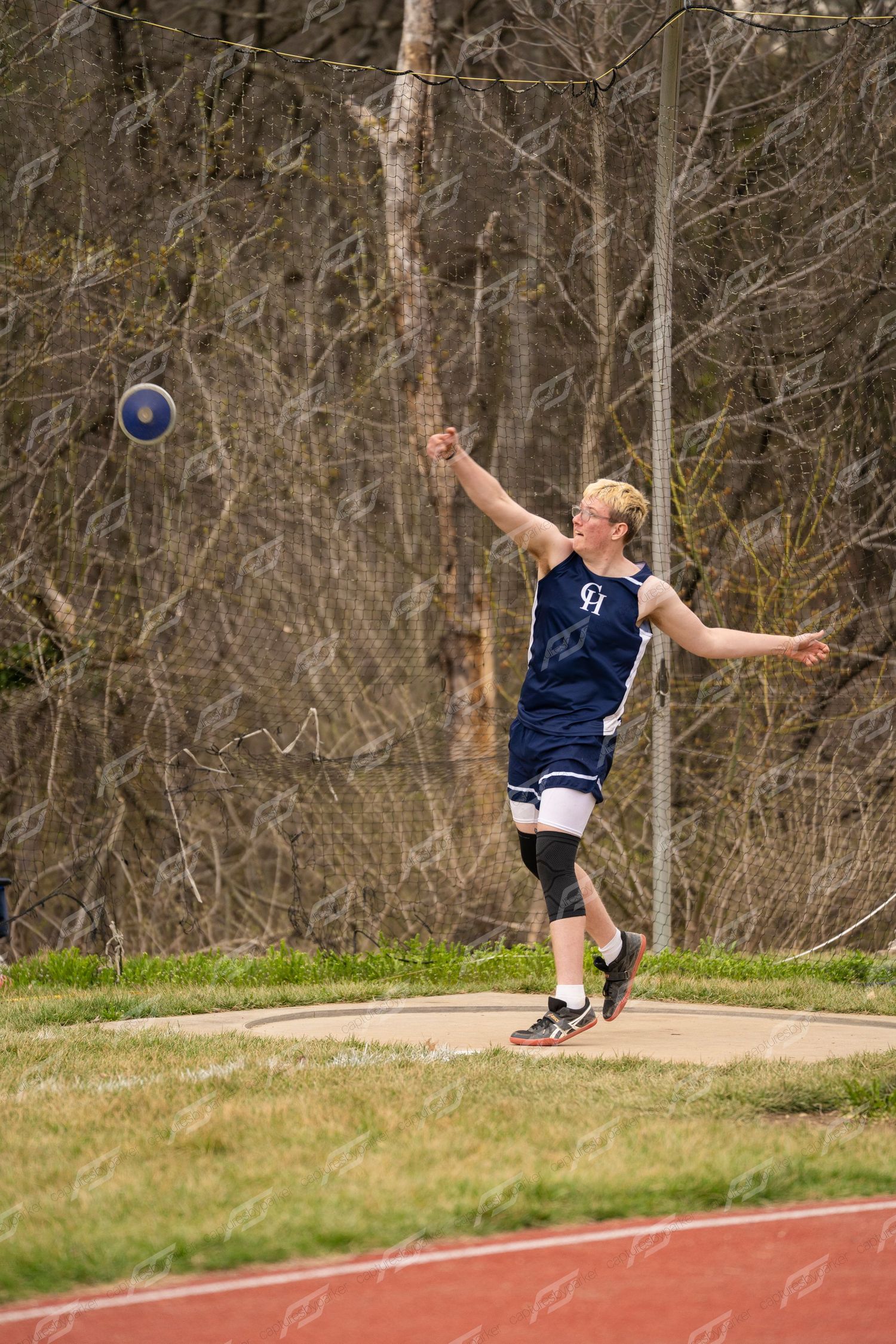 A high school athlete releases a discus during an outdoor track and field competition on a red track.
