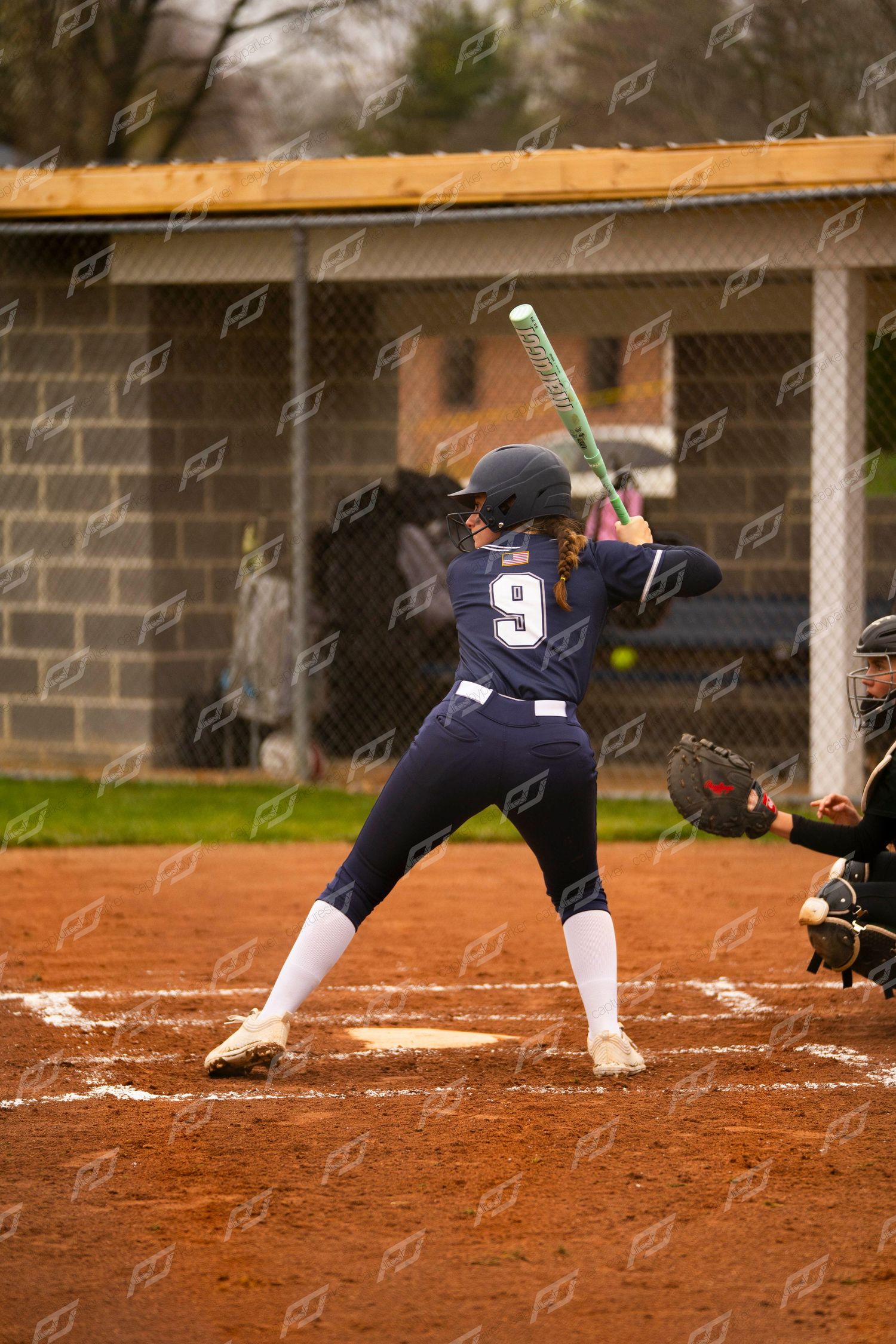 Softball player number 9 in navy uniform stands at bat awaiting a pitch with catcher behind her.