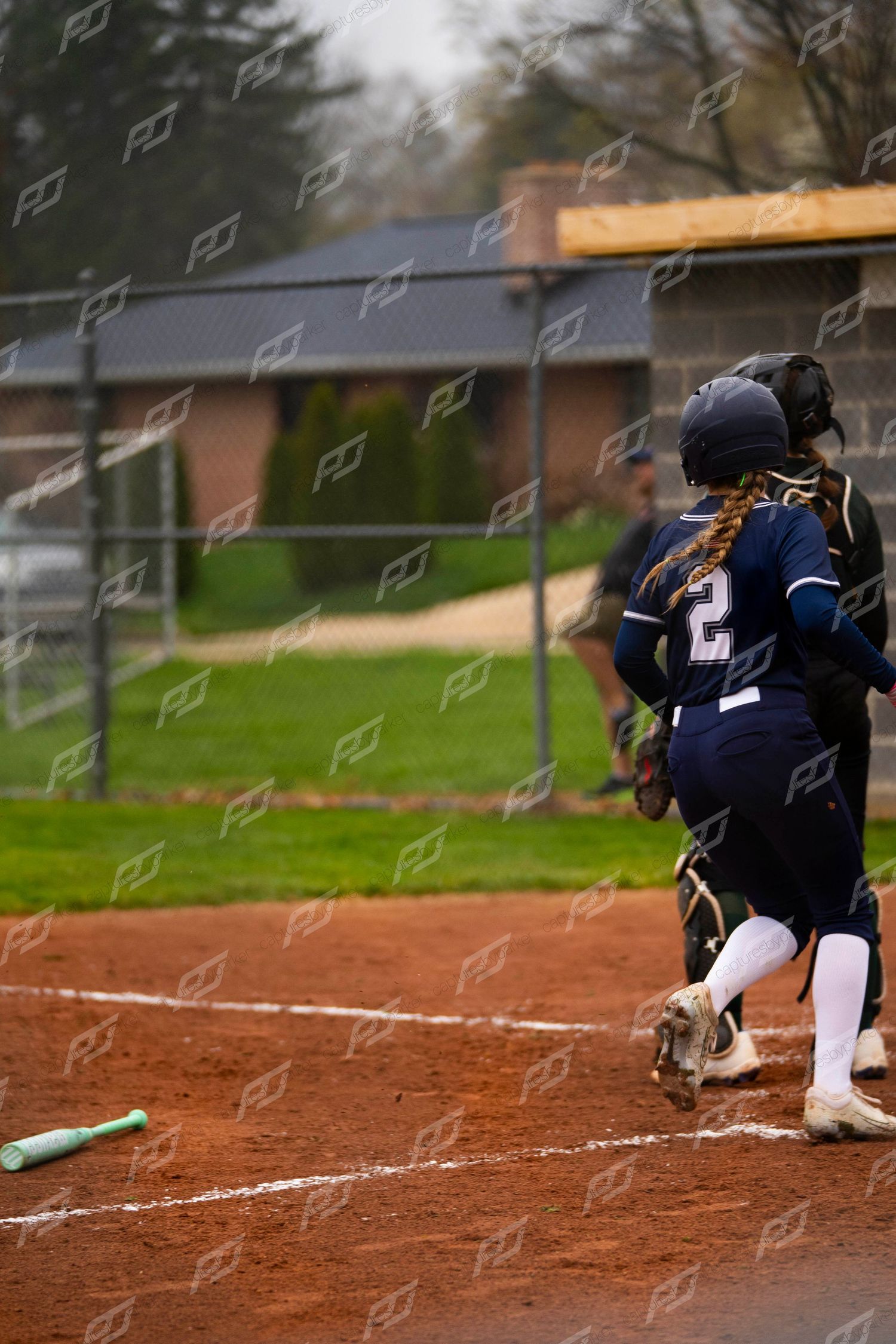 Female softball player #2 with orange braid runs to first base after dropping the bat during a game.