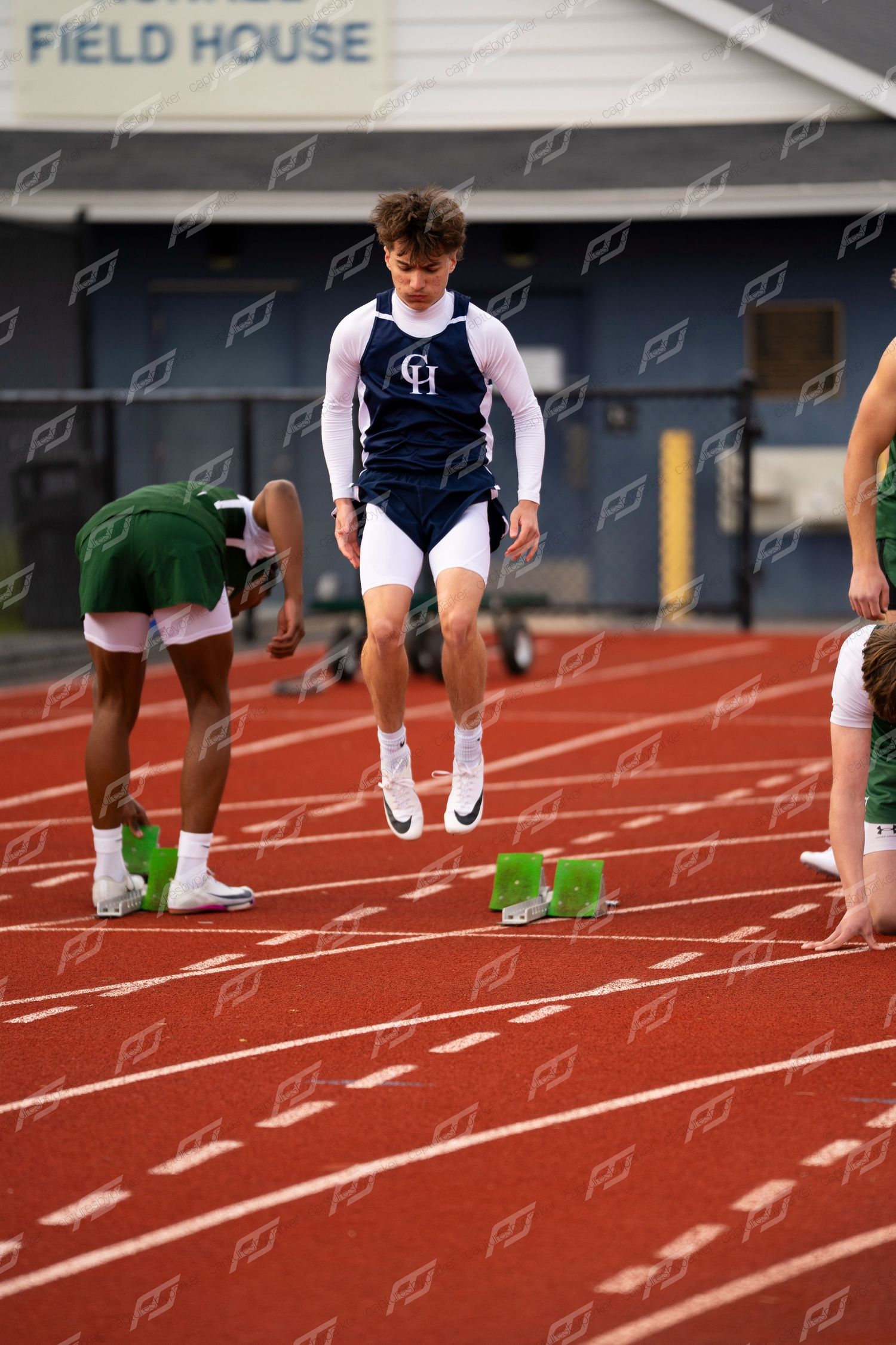 Male sprinter in white and navy CH uniform jumps at starting blocks on red outdoor track.