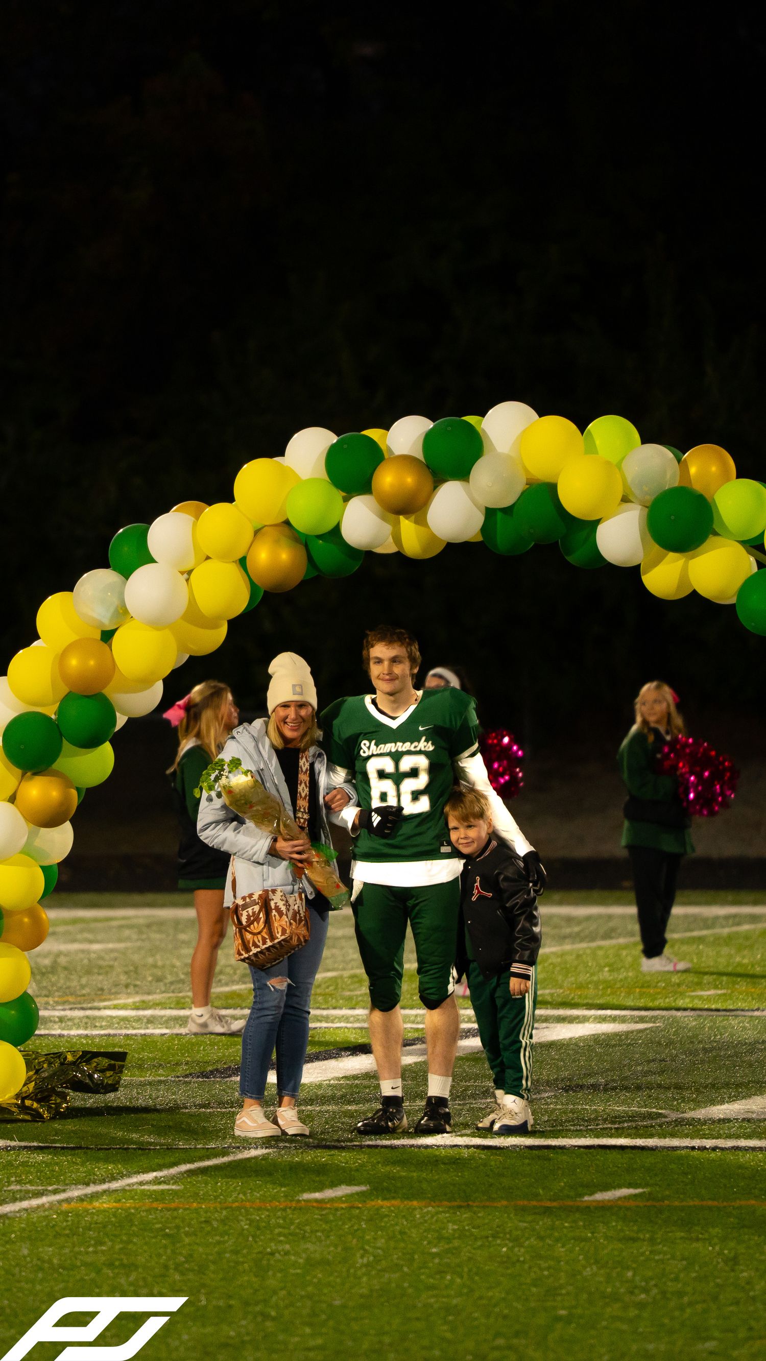Shamrocks football player #62 celebrates senior night with two family members under colorful balloon arch.