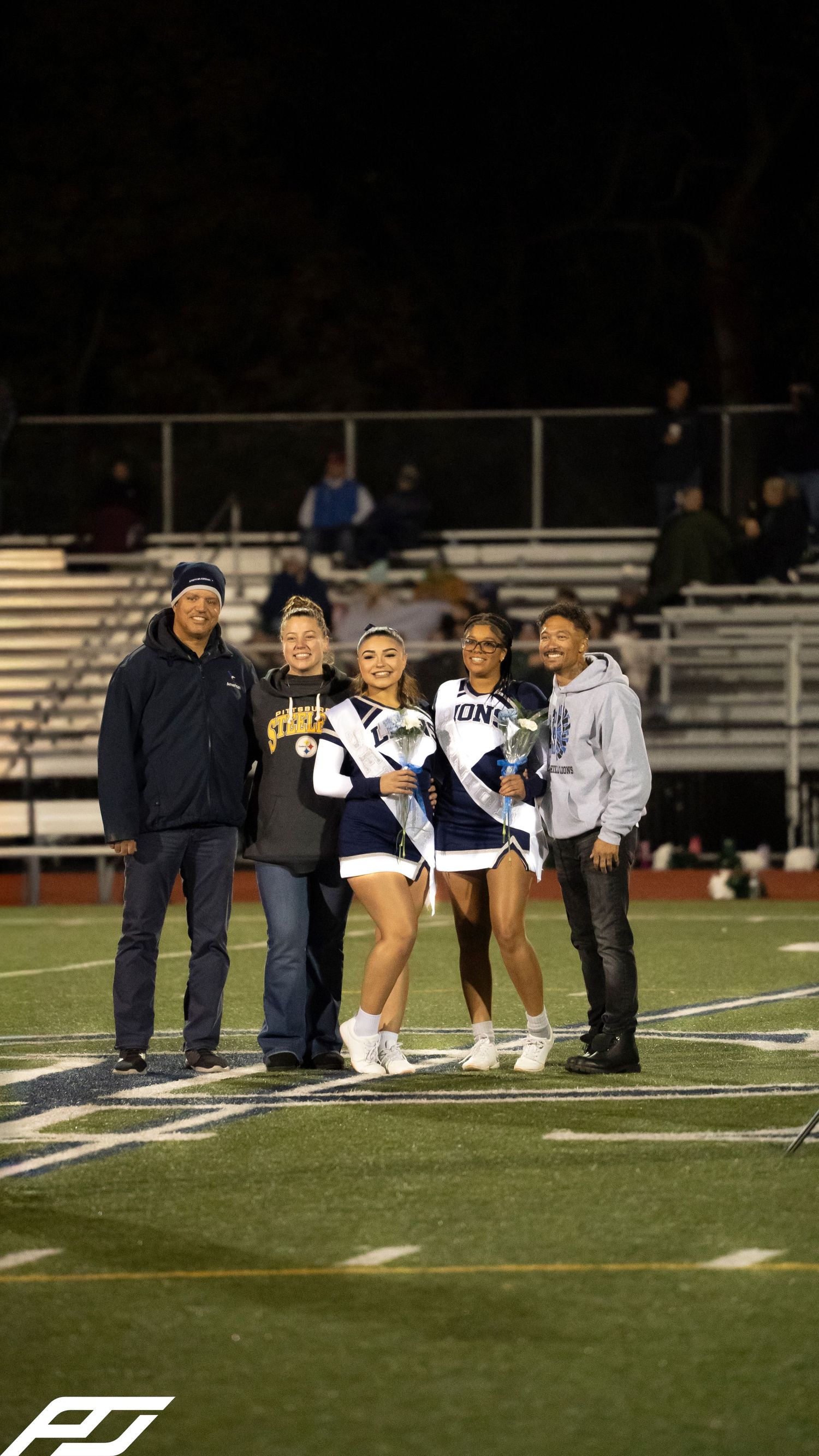 Two cheerleaders in blue and white uniforms pose with family members on a football field at night.