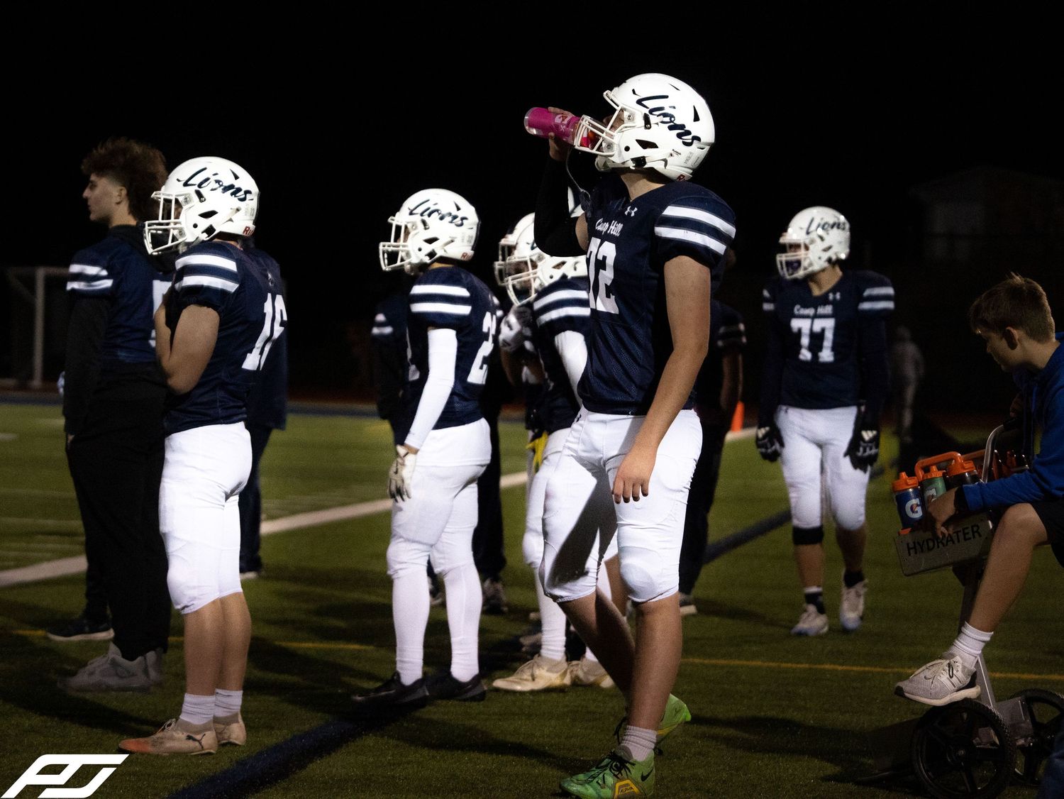 Camp Hill Lions youth football players stand on the sideline during a night game.