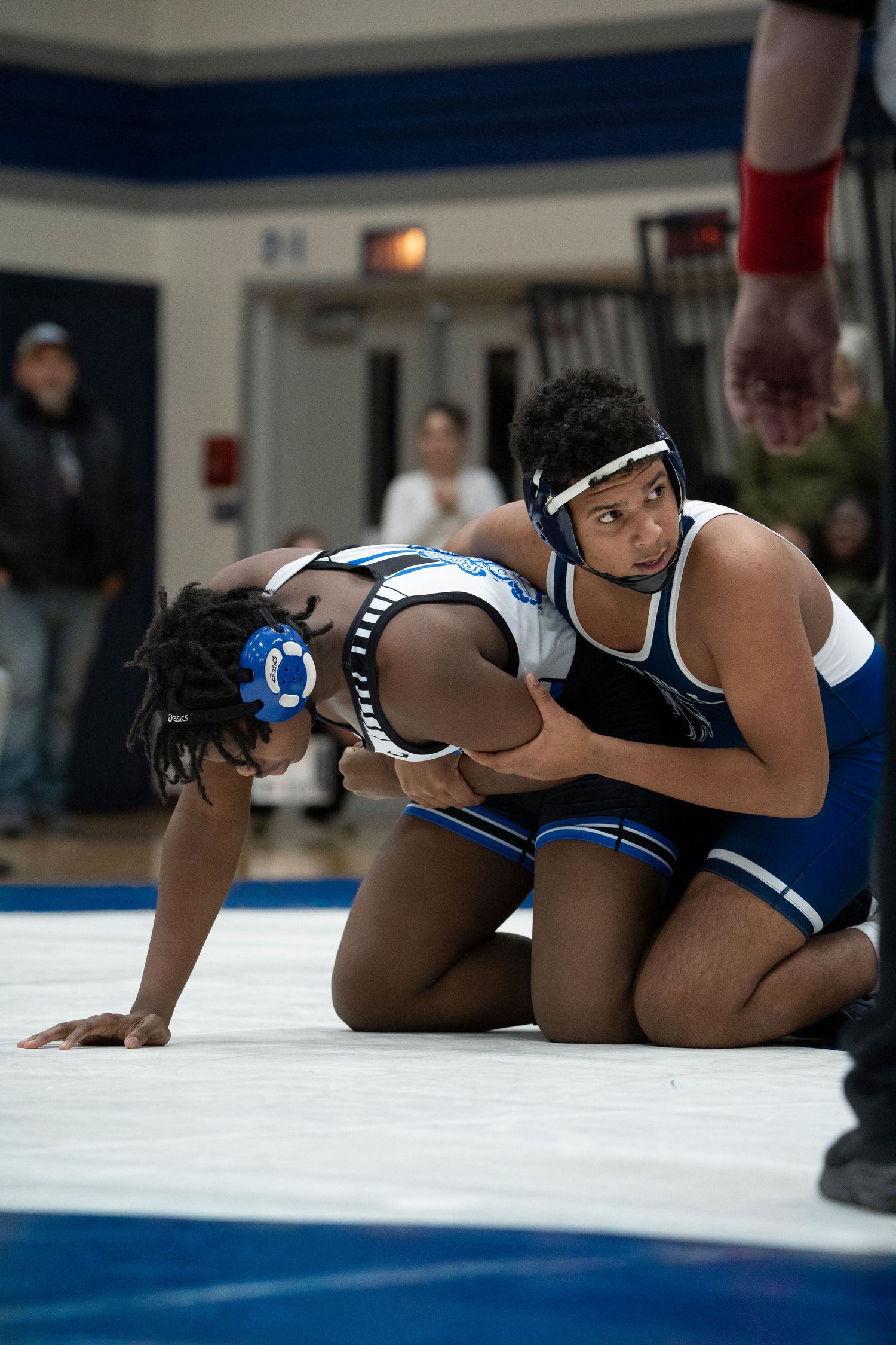 A wrestler in navy blue controls his opponent on the mat during a competitive high school wrestling match.