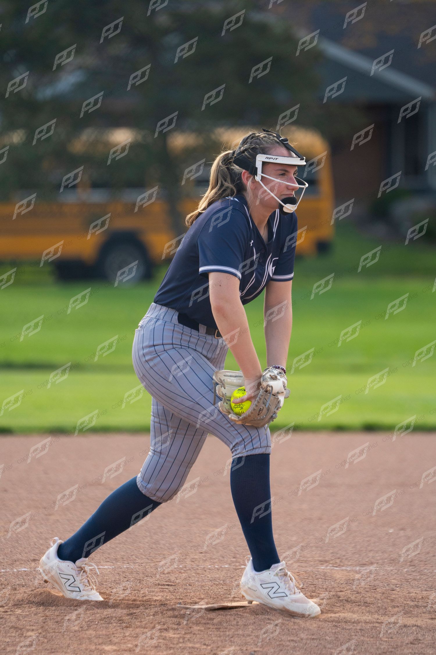 Female softball pitcher in navy uniform winds up to throw yellow softball.