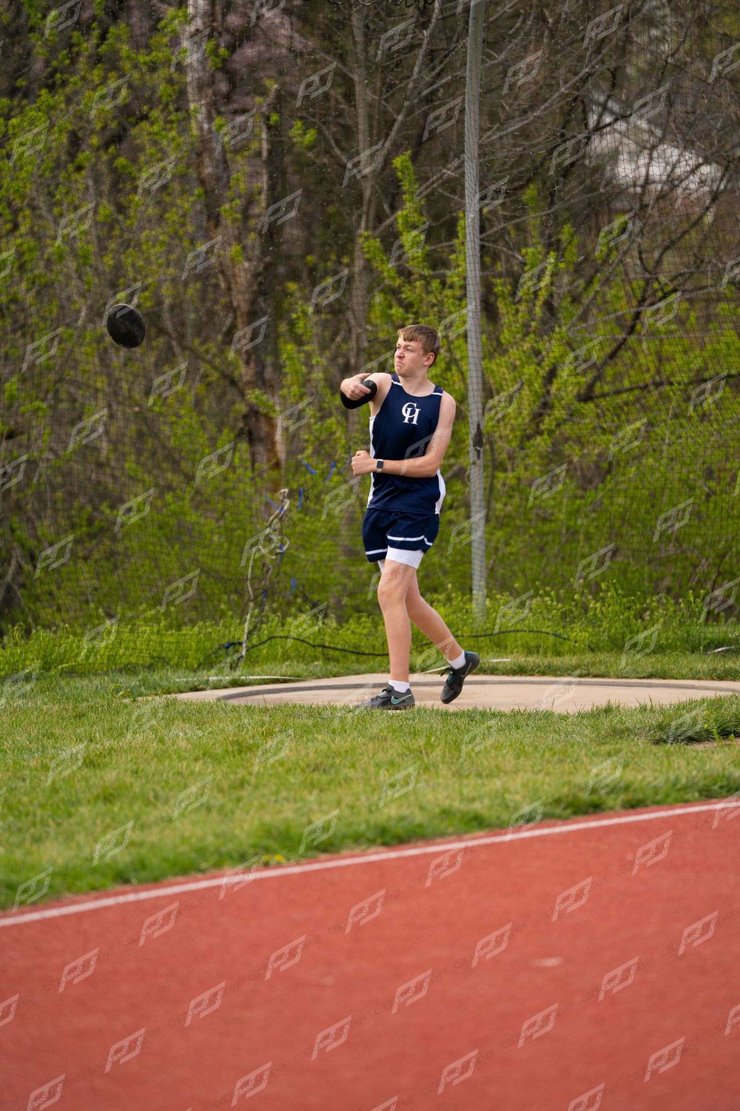 Male athlete in navy uniform throws a shot put during an outdoor track and field competition.