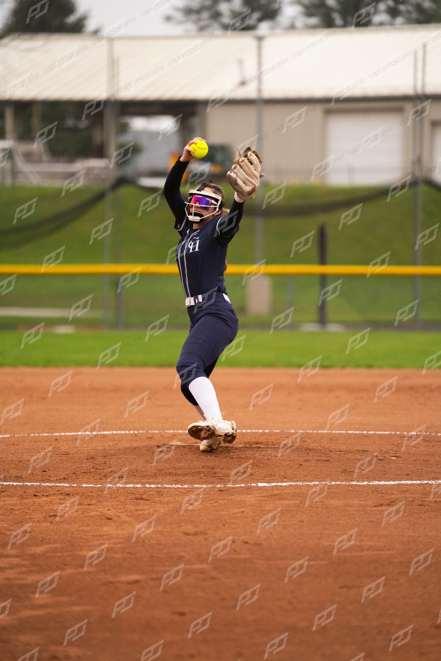 Softball pitcher in navy uniform mid-windup delivering an underhand pitch on a clay infield.