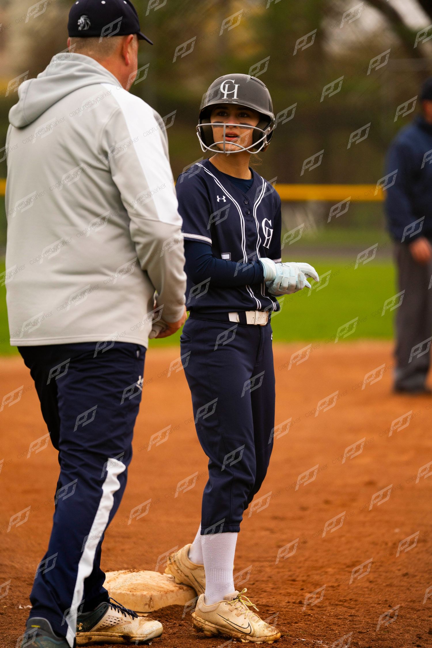Softball player in navy uniform talks with coach at first base during a game.