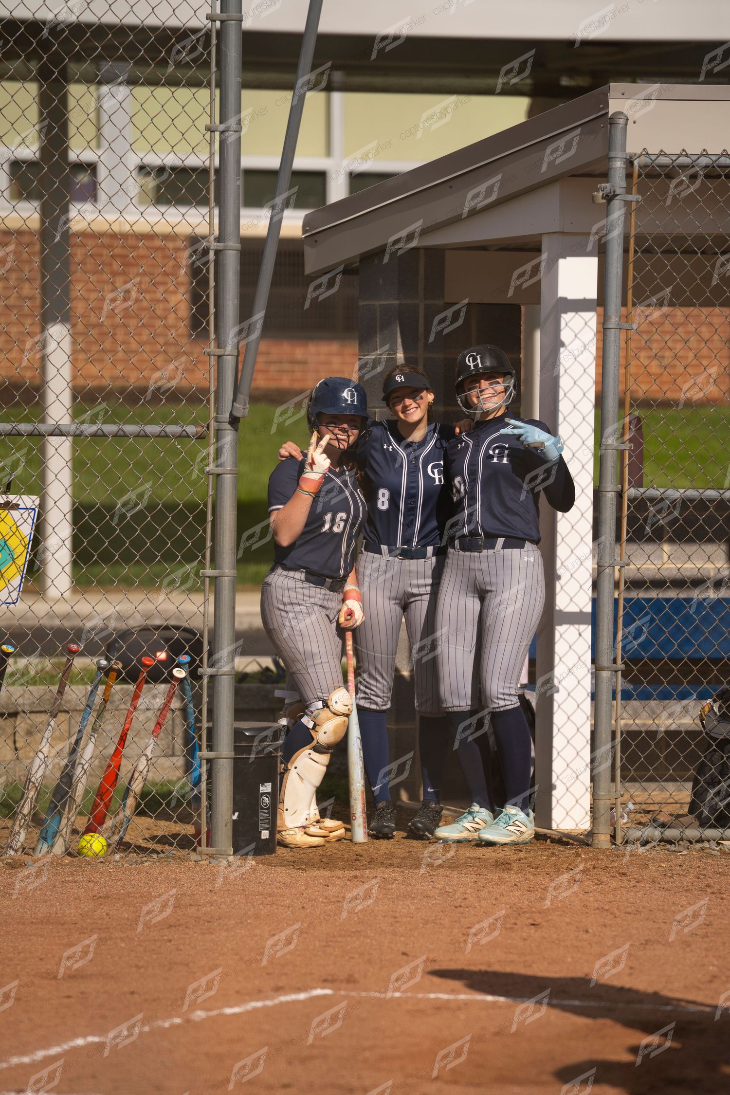 Three female softball players in navy uniforms pose together near the dugout entrance during a game.
