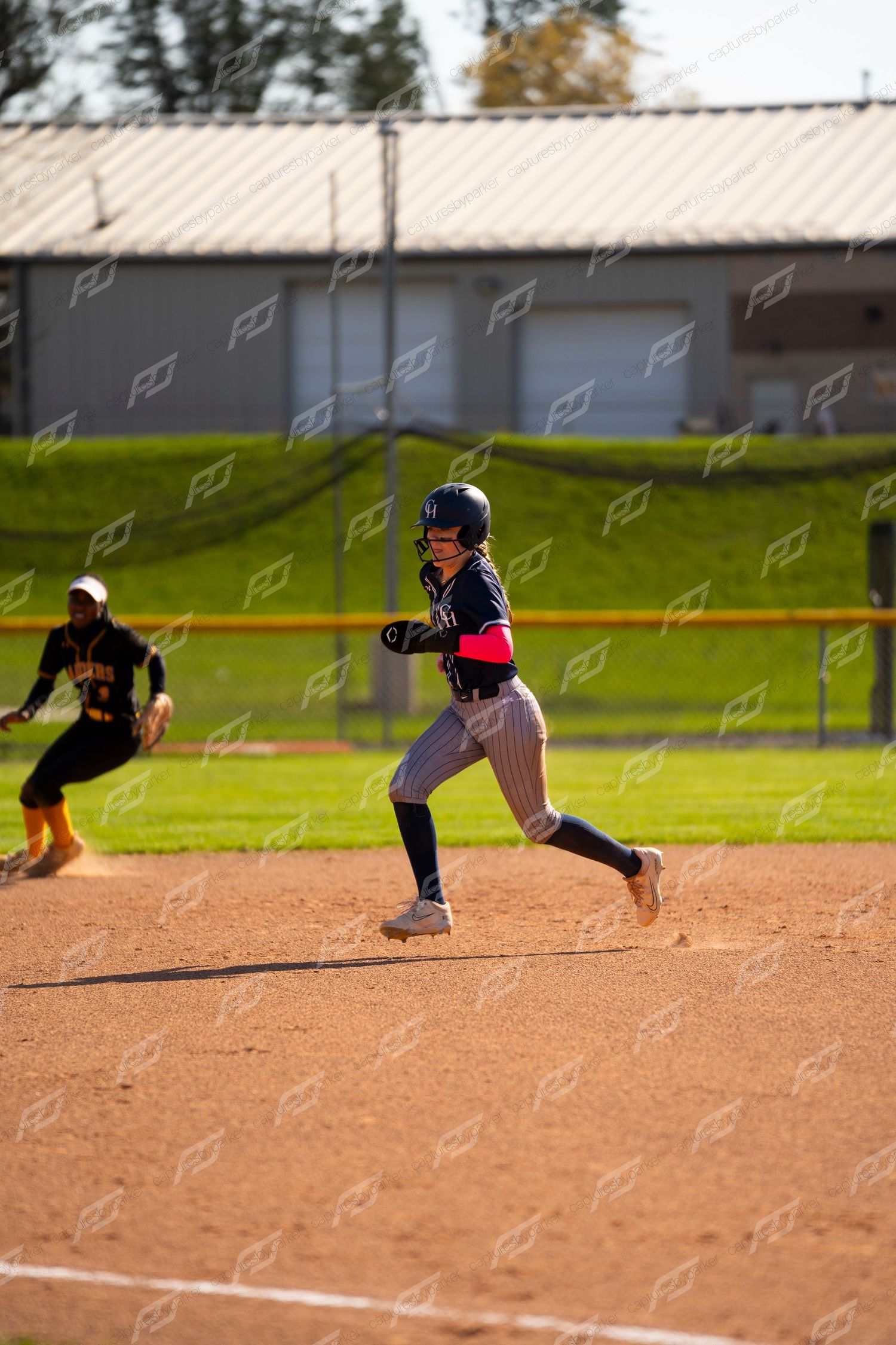 Female softball player in navy uniform rounds the bases at full sprint during a competitive game.