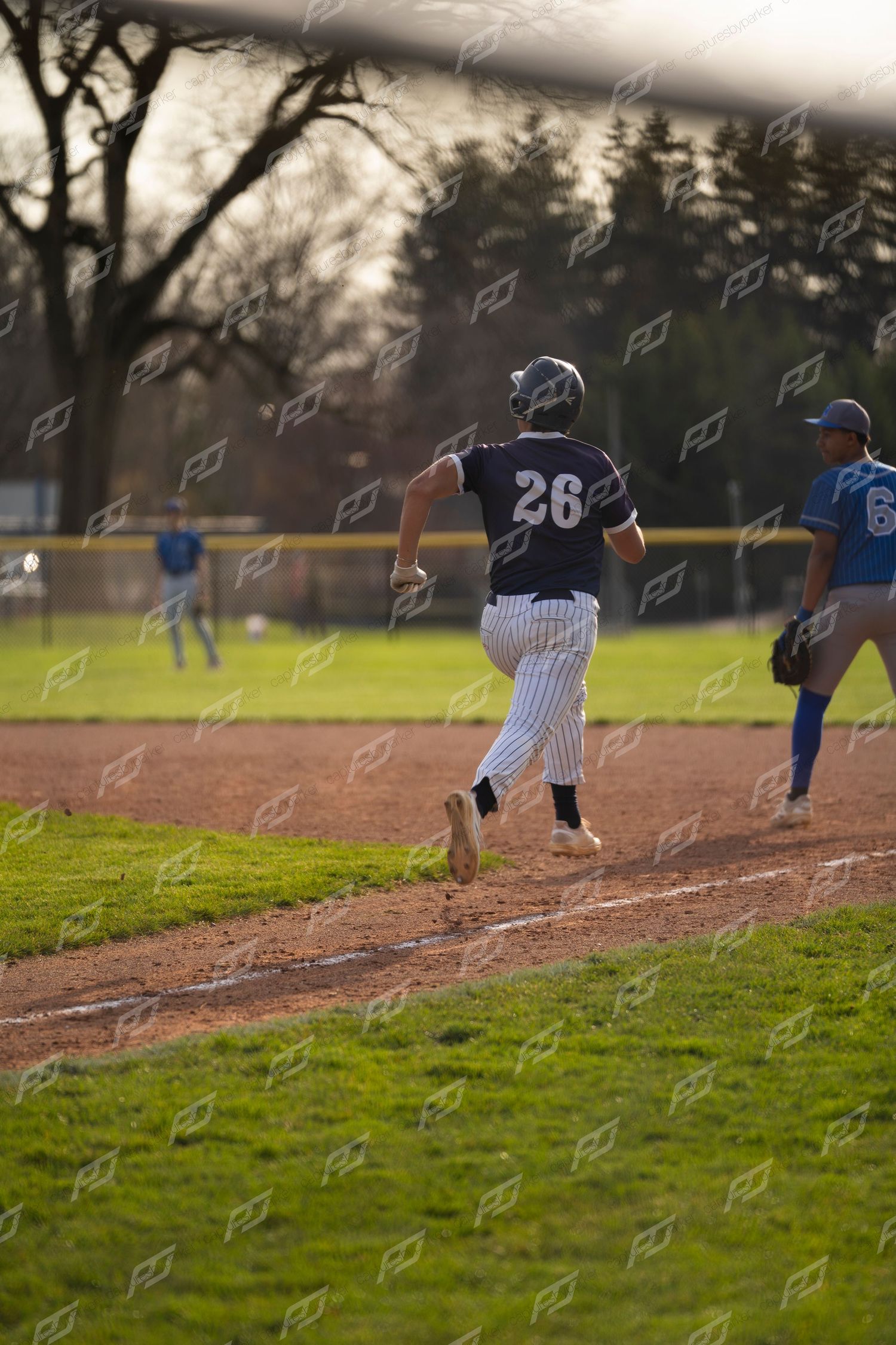 Baseball player #26 in dark uniform runs the base path during a youth baseball game on a sunny day.
