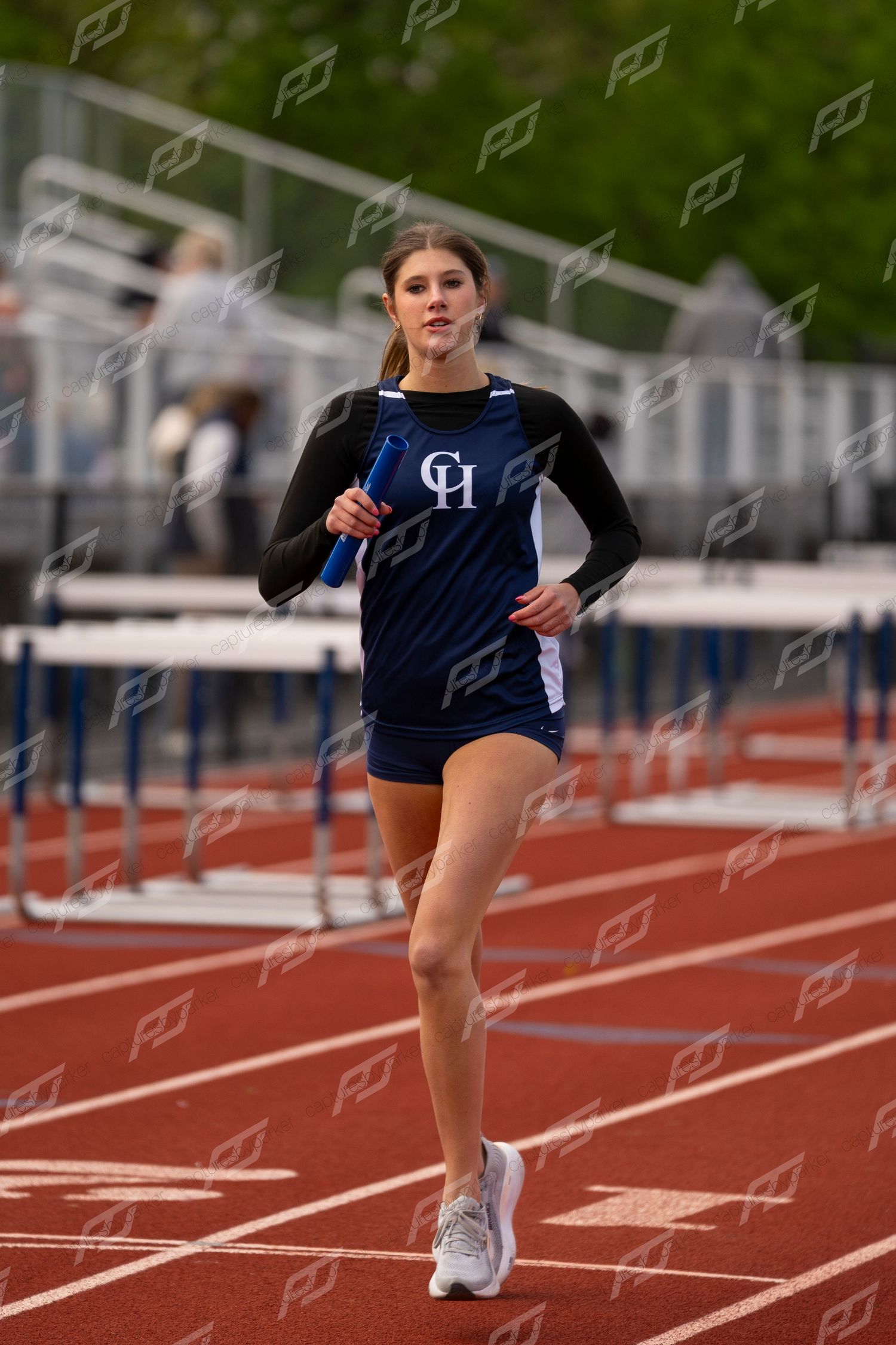 Female CH track athlete in navy uniform running a relay race while holding a blue baton near hurdles.