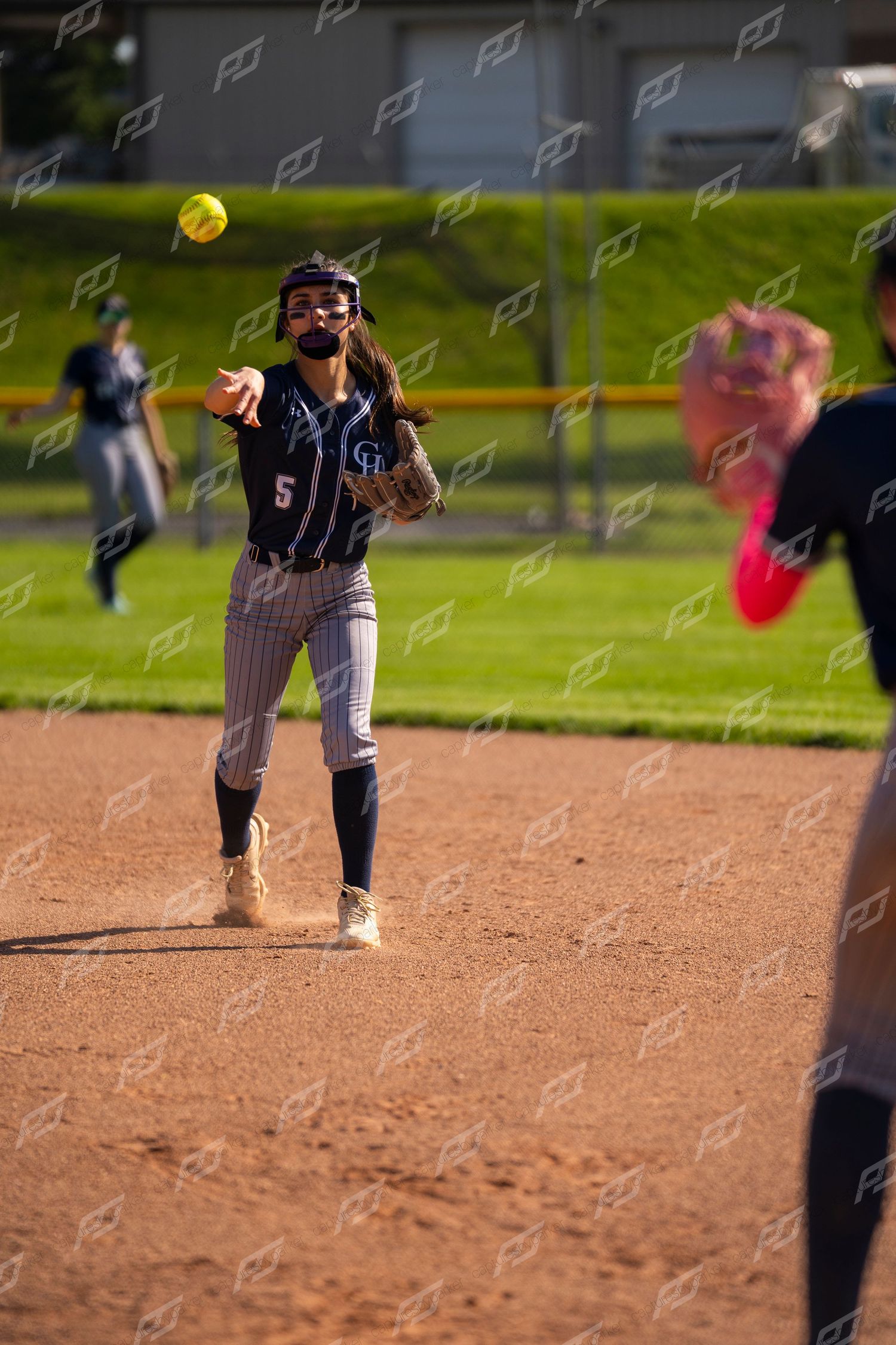 Softball player #5 throws to first base during an outdoor game on a sunny day.