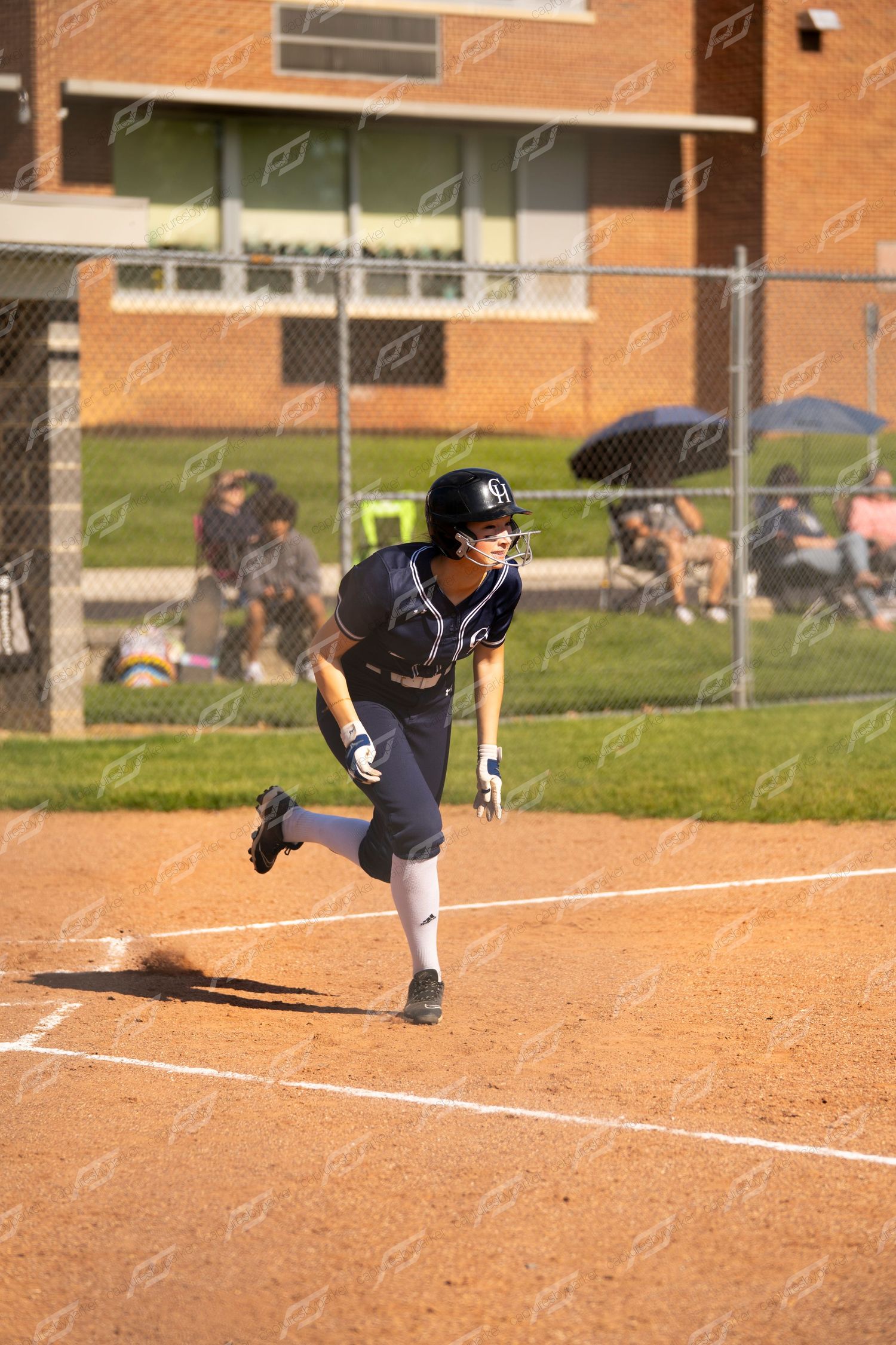 Softball player in navy uniform sprinting from home plate during a sunny afternoon game.