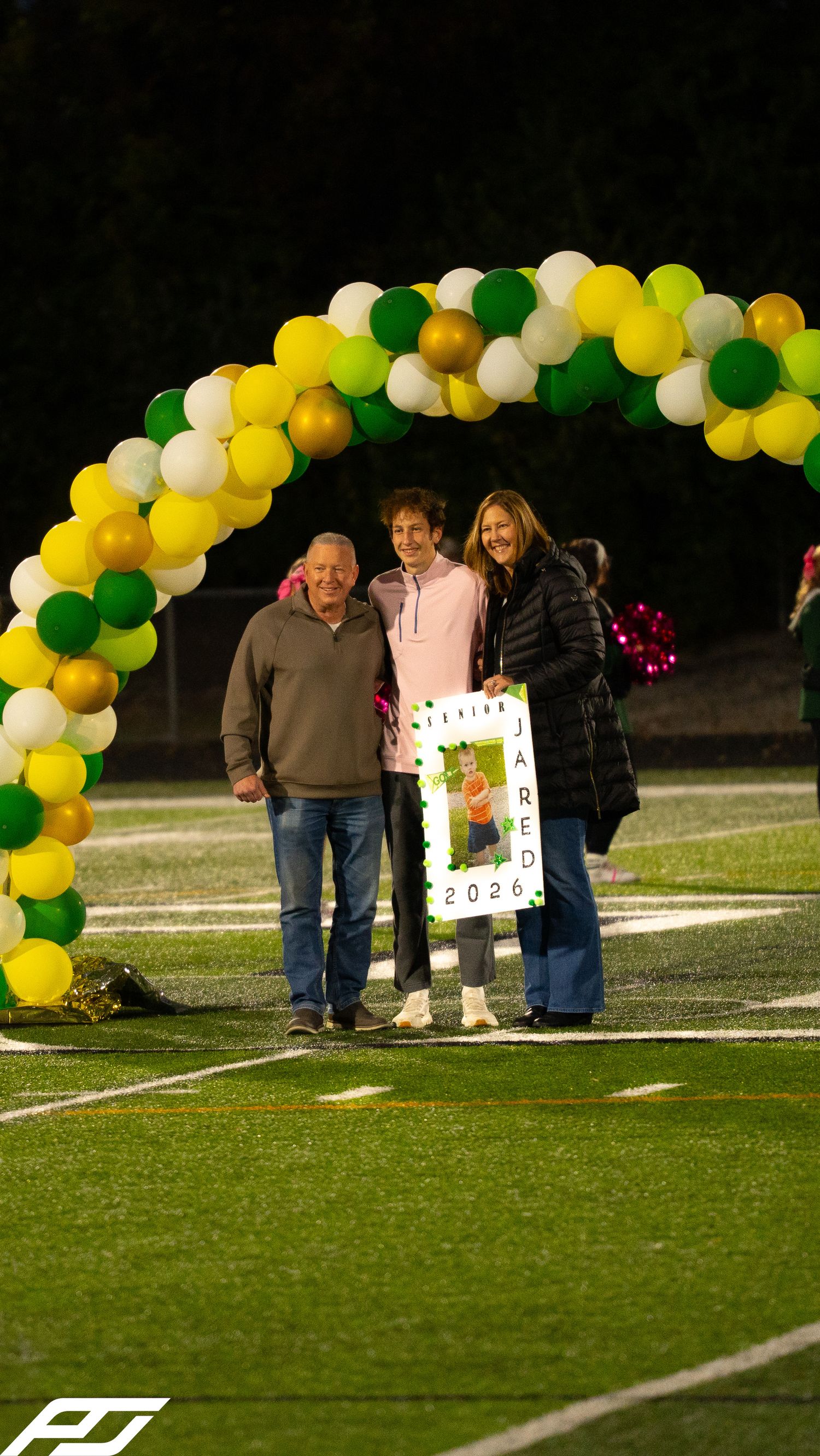 Jared 2026 senior night recognition with parents under green and gold balloon arch on football field.