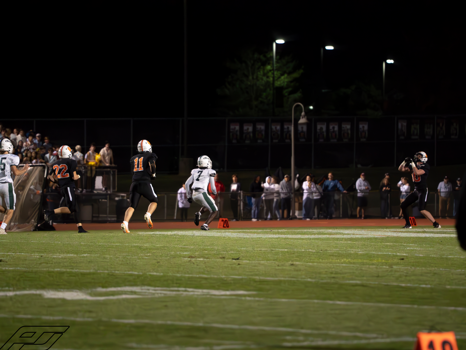 Football players in orange uniforms run a play near the sideline with fans watching under bright stadium lights.