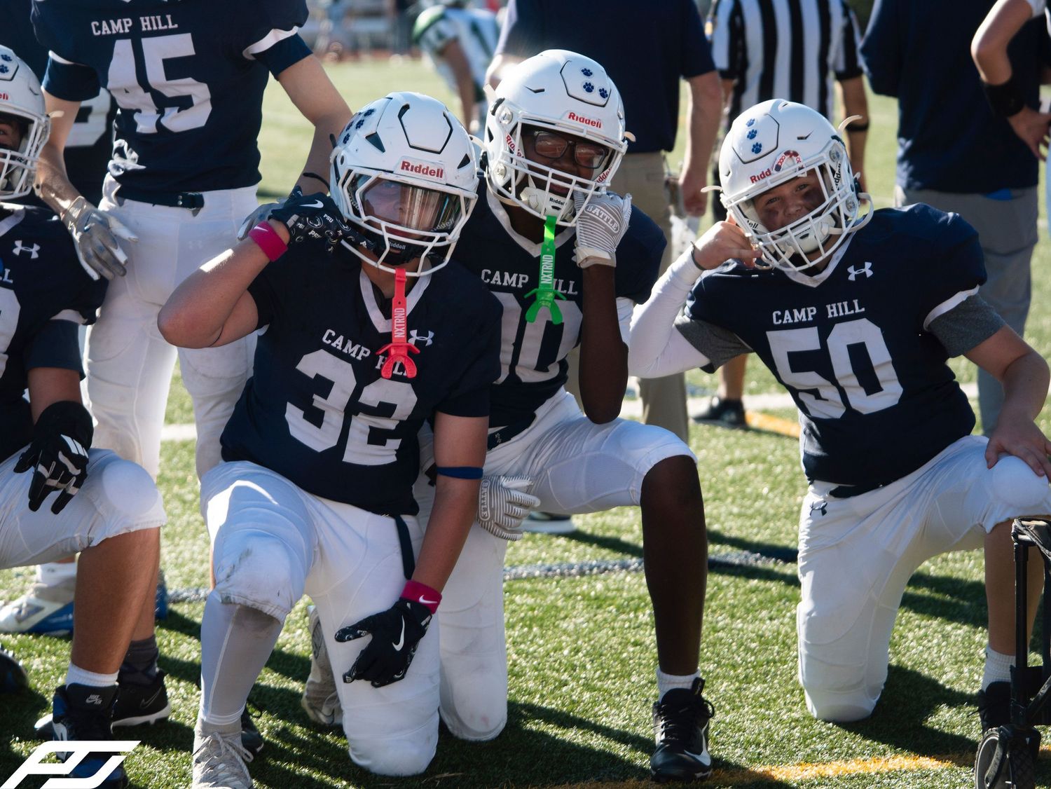 Three youth football players in Camp Hill uniforms kneel on the field during a game.