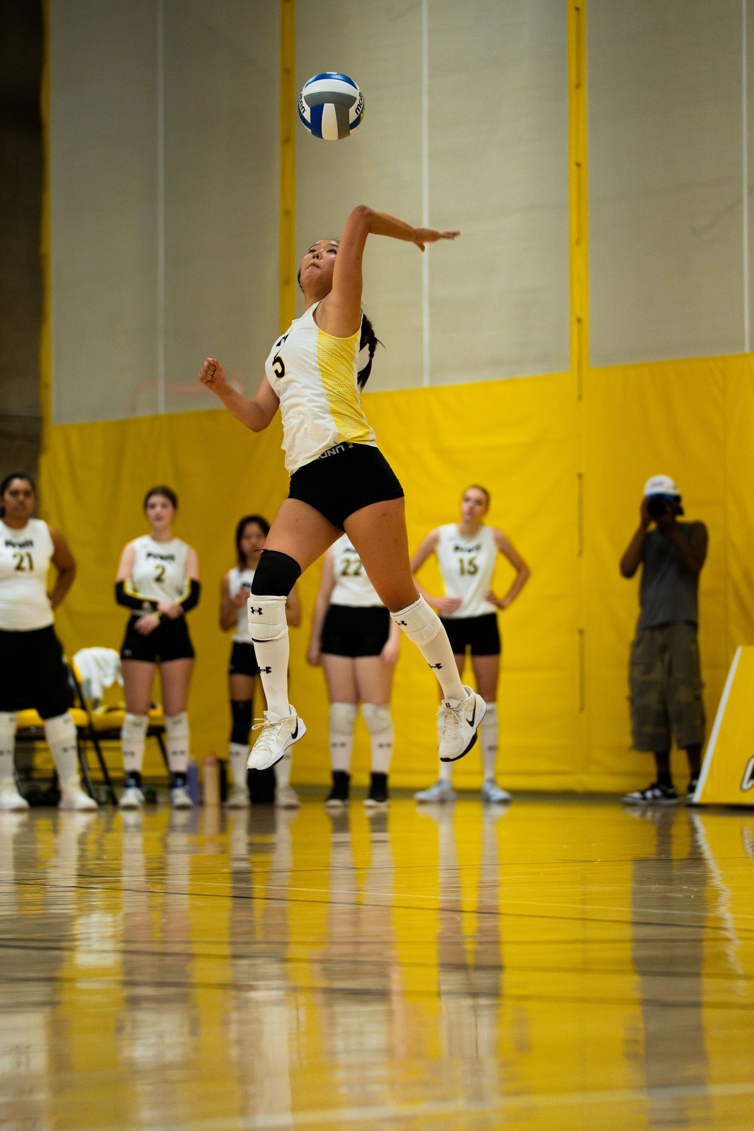 Female volleyball player jumps to serve ball during indoor match, teammates watching in background.