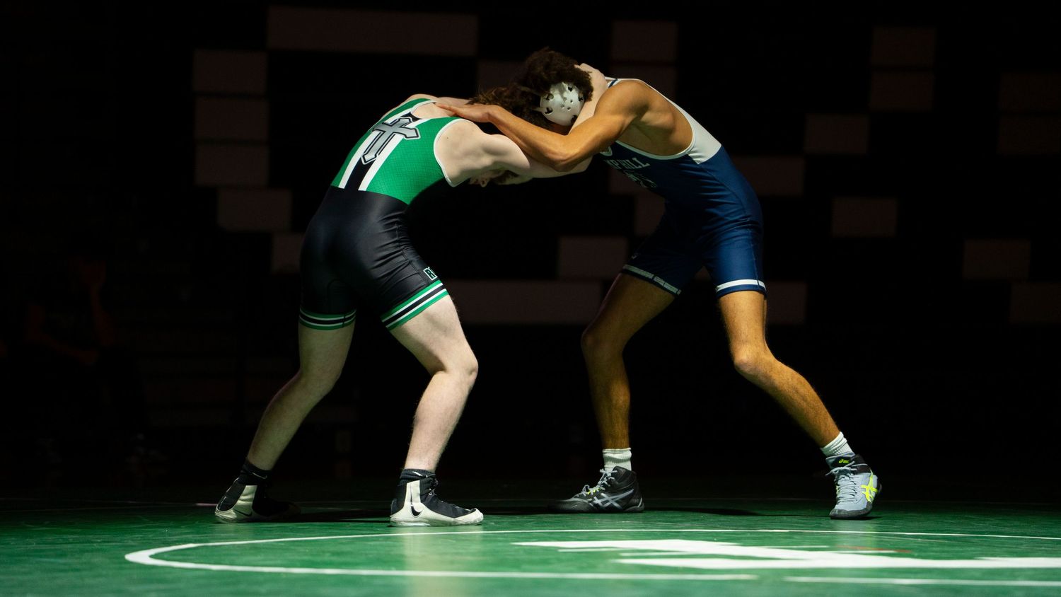 Two wrestlers in green and navy singlets compete on a green mat in a dimly lit gymnasium.