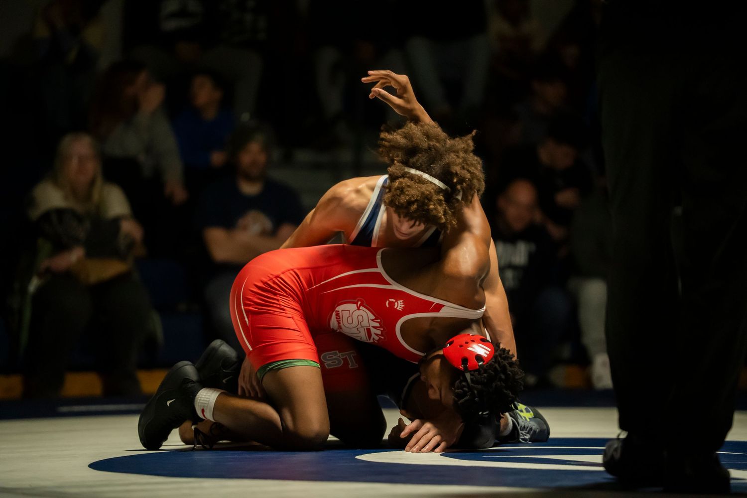 Wrestler in red singlet controls opponent on the mat during a competitive wrestling match.