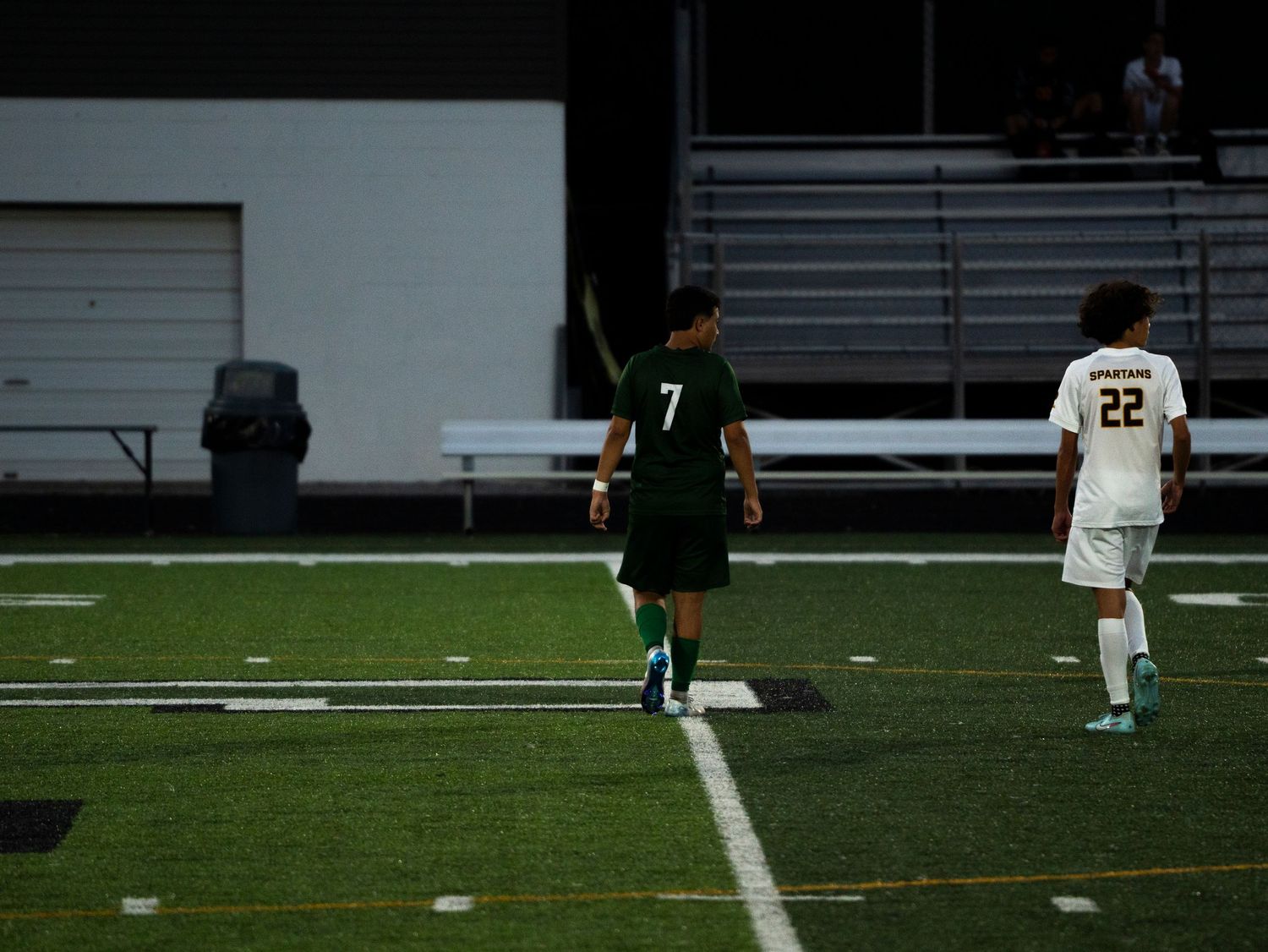 Trinity soccer player #7 walks along sideline near opponent #22 during evening match on turf field.